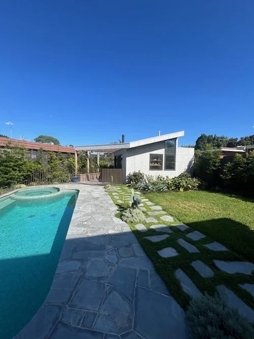 Modern backyard with a swimming pool, stone pathway, grassy lawn, and contemporary house under a clear blue sky.