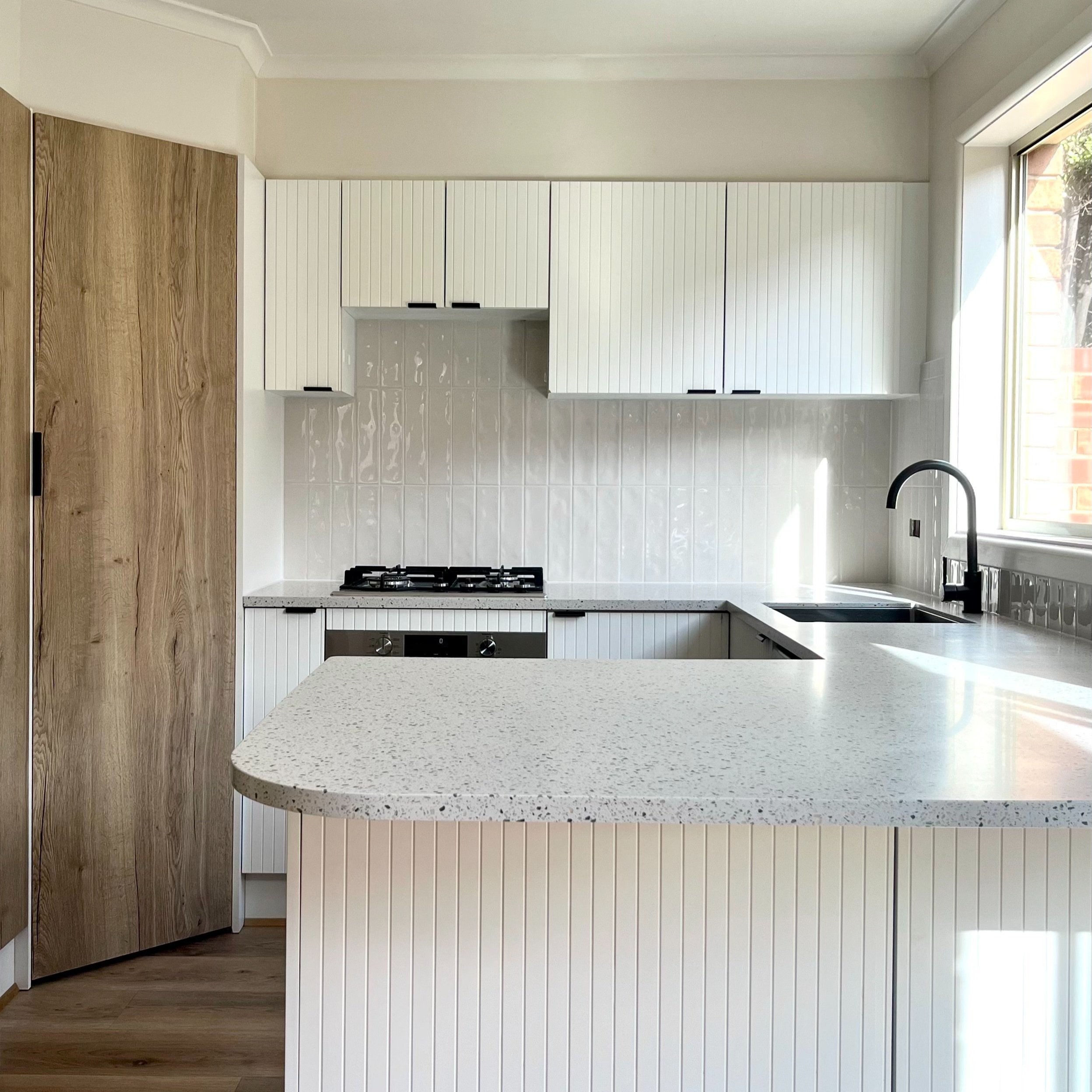 Modern kitchen with white cabinetry, wood accents, quartz countertop, and stainless steel appliances, featuring a black faucet and a large window.
