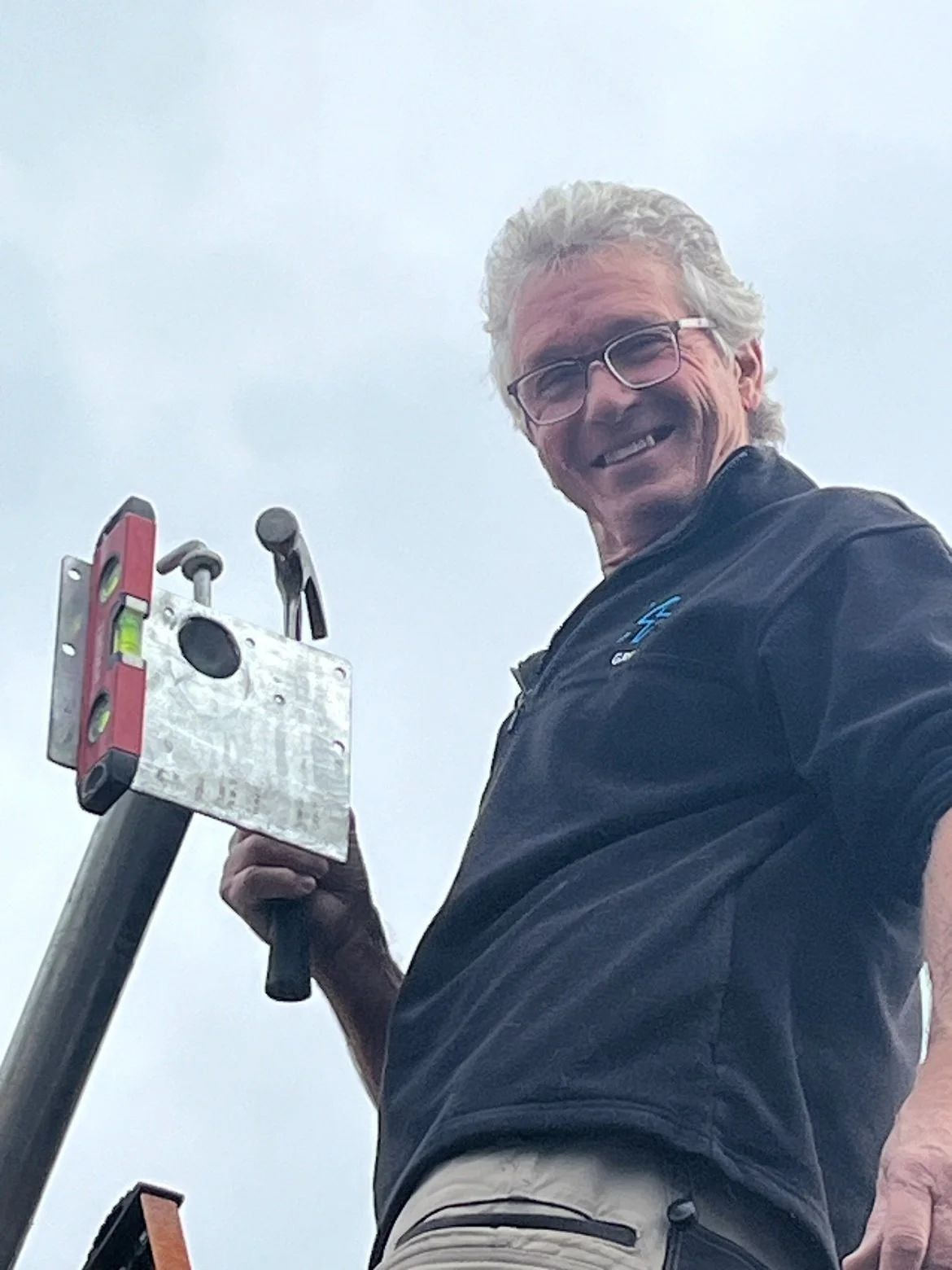Person smiling while holding a hammer and standing next to a mounted level tool against a backdrop of a cloudy sky.