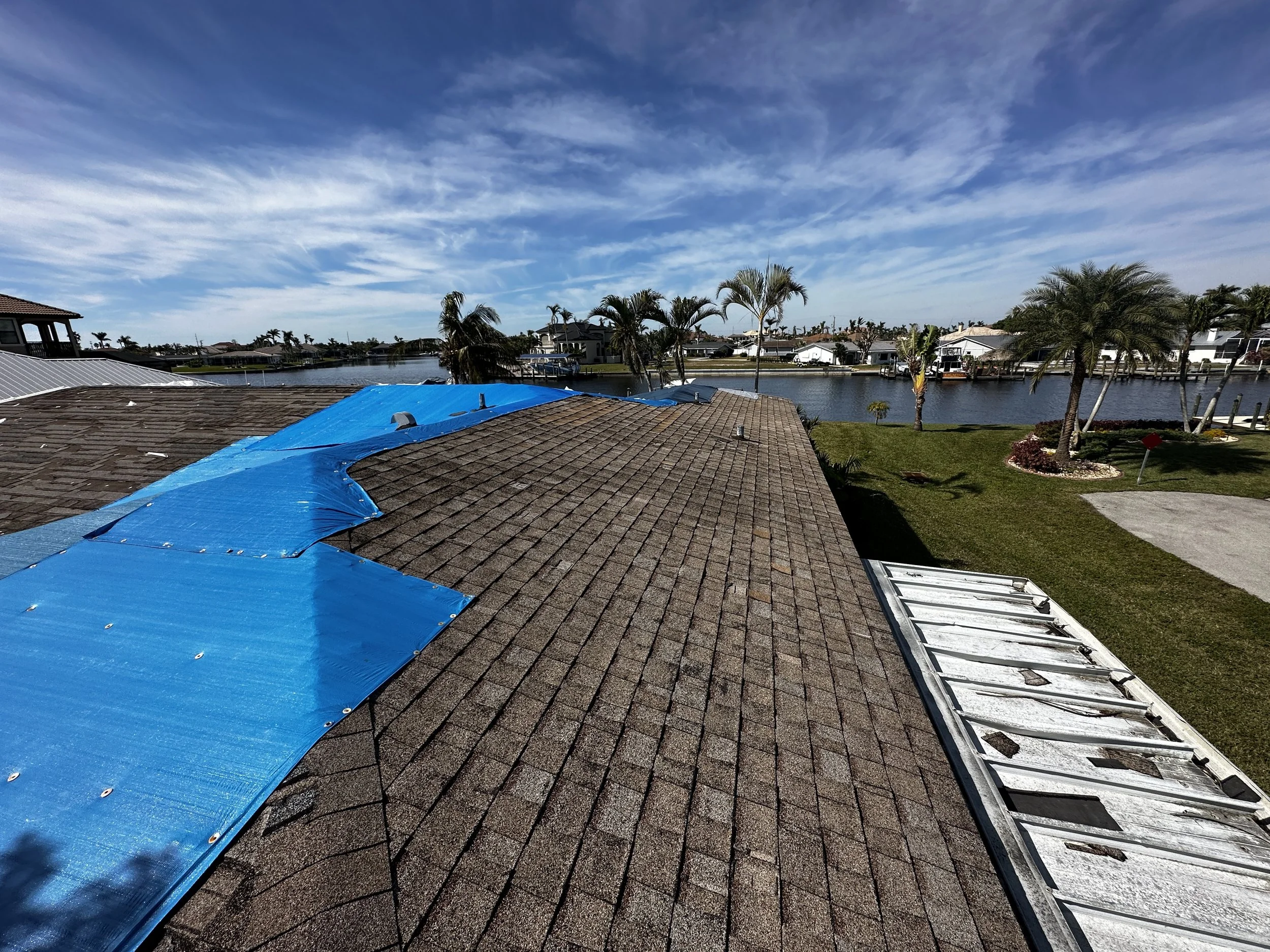 Residential house roof with blue tarpaulin covering part of it, near a canal with palm trees and other houses in the background, under a partly cloudy sky.