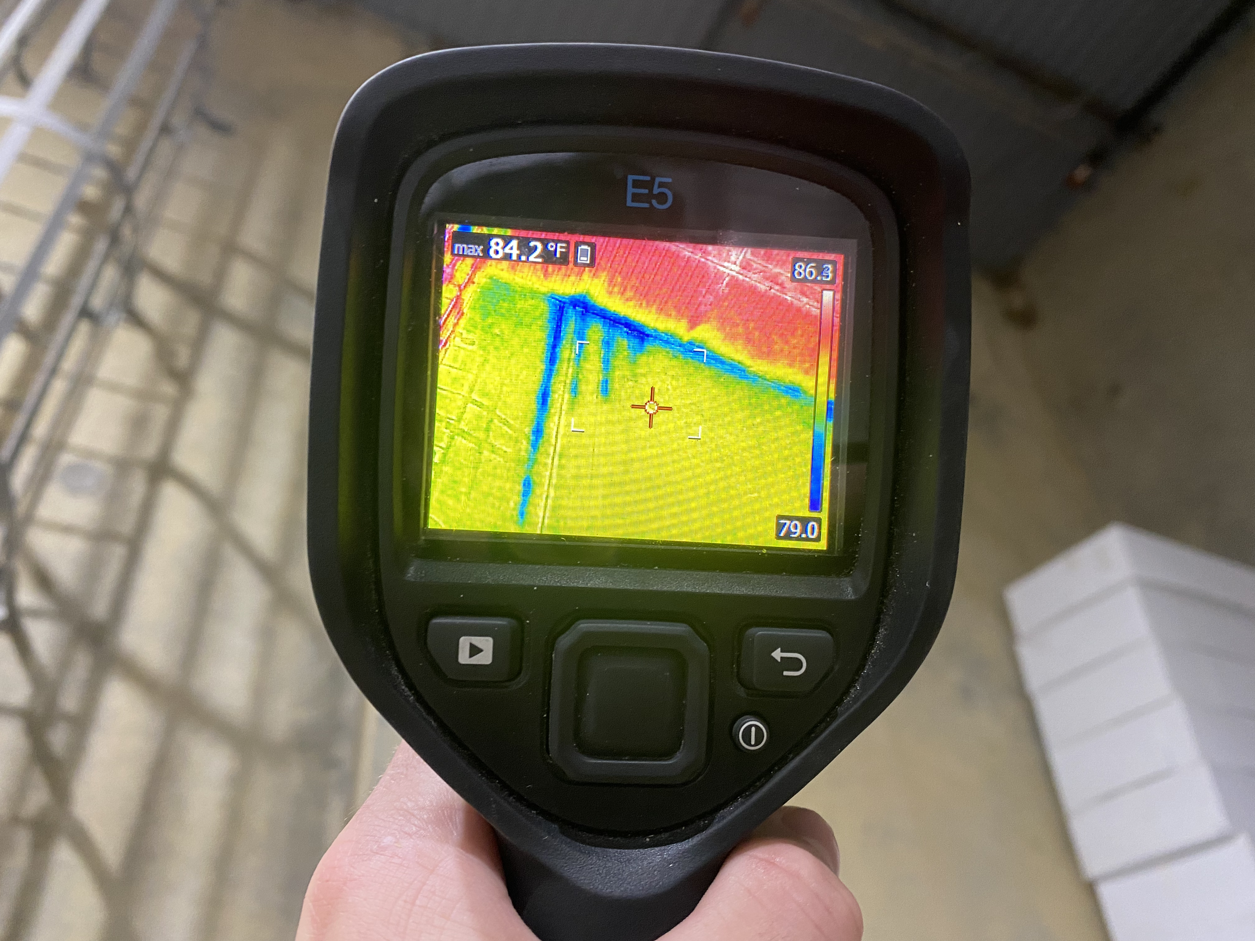 Hand holding an infrared thermal camera showing a thermal image of a wall with hot and cold spots in a warehouse.