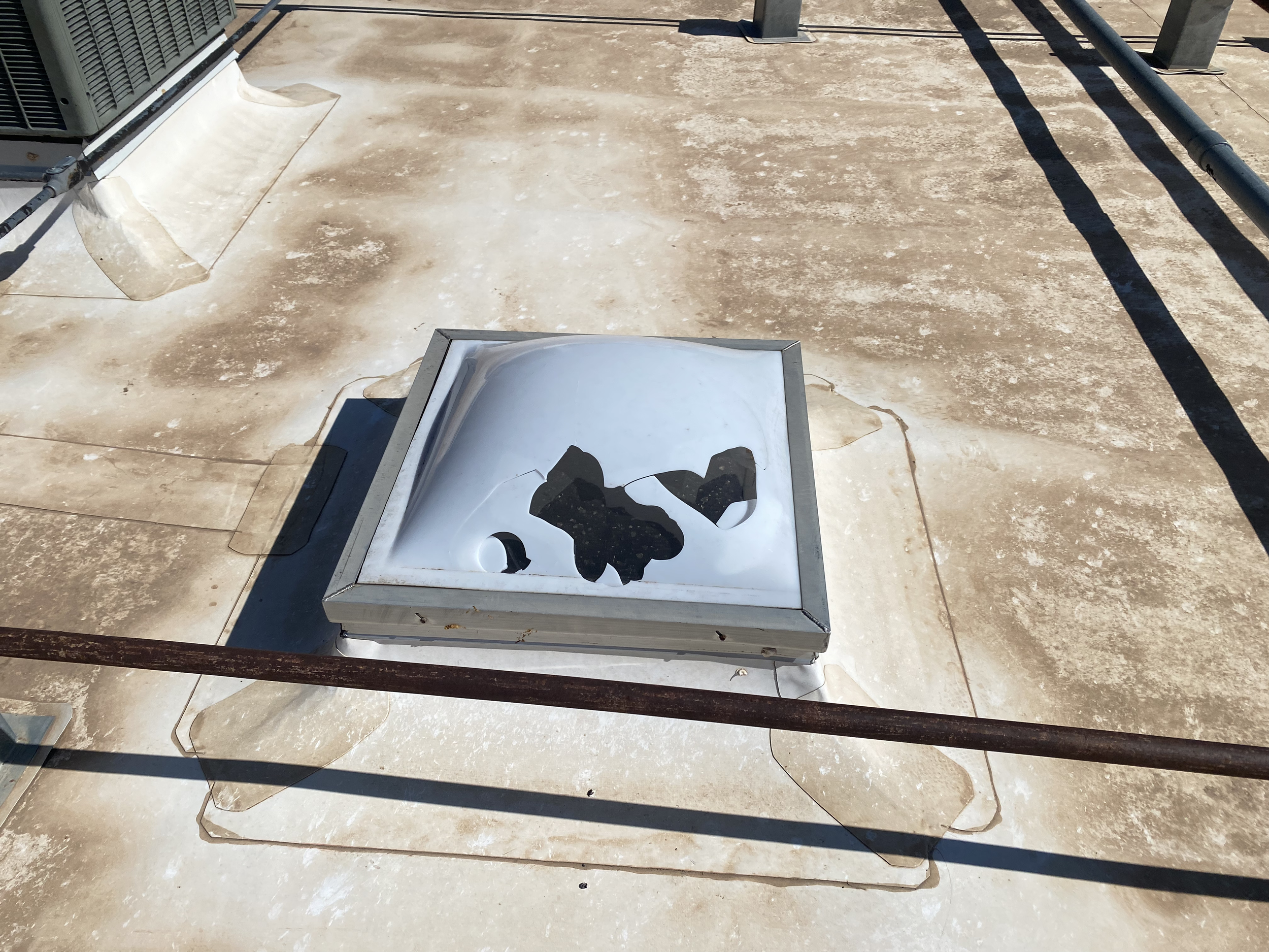 A hail damaged rooftop skylight with broken glass and a partially torn protective film, on a TPO commercial flat roof