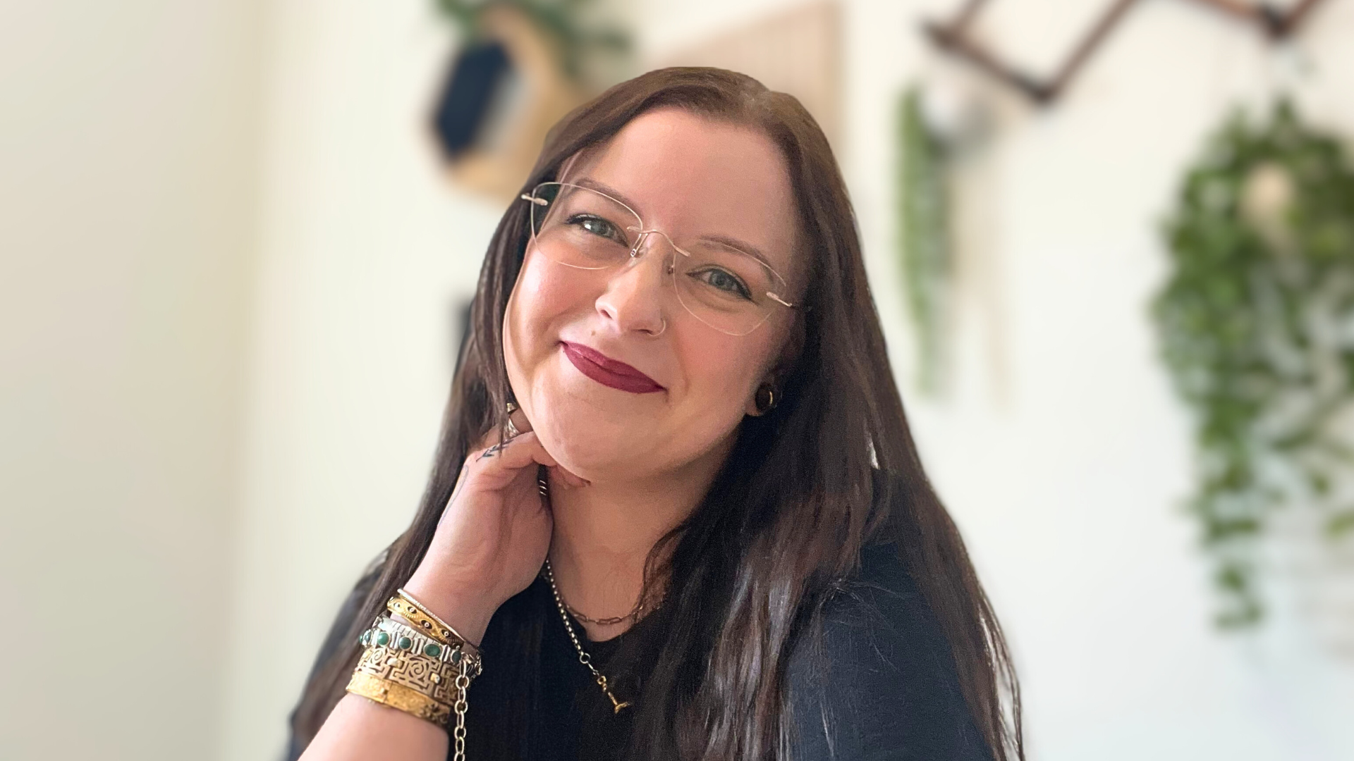 Smiling person with glasses, long dark hair, wearing jewelry, indoors with blurred plants in background.