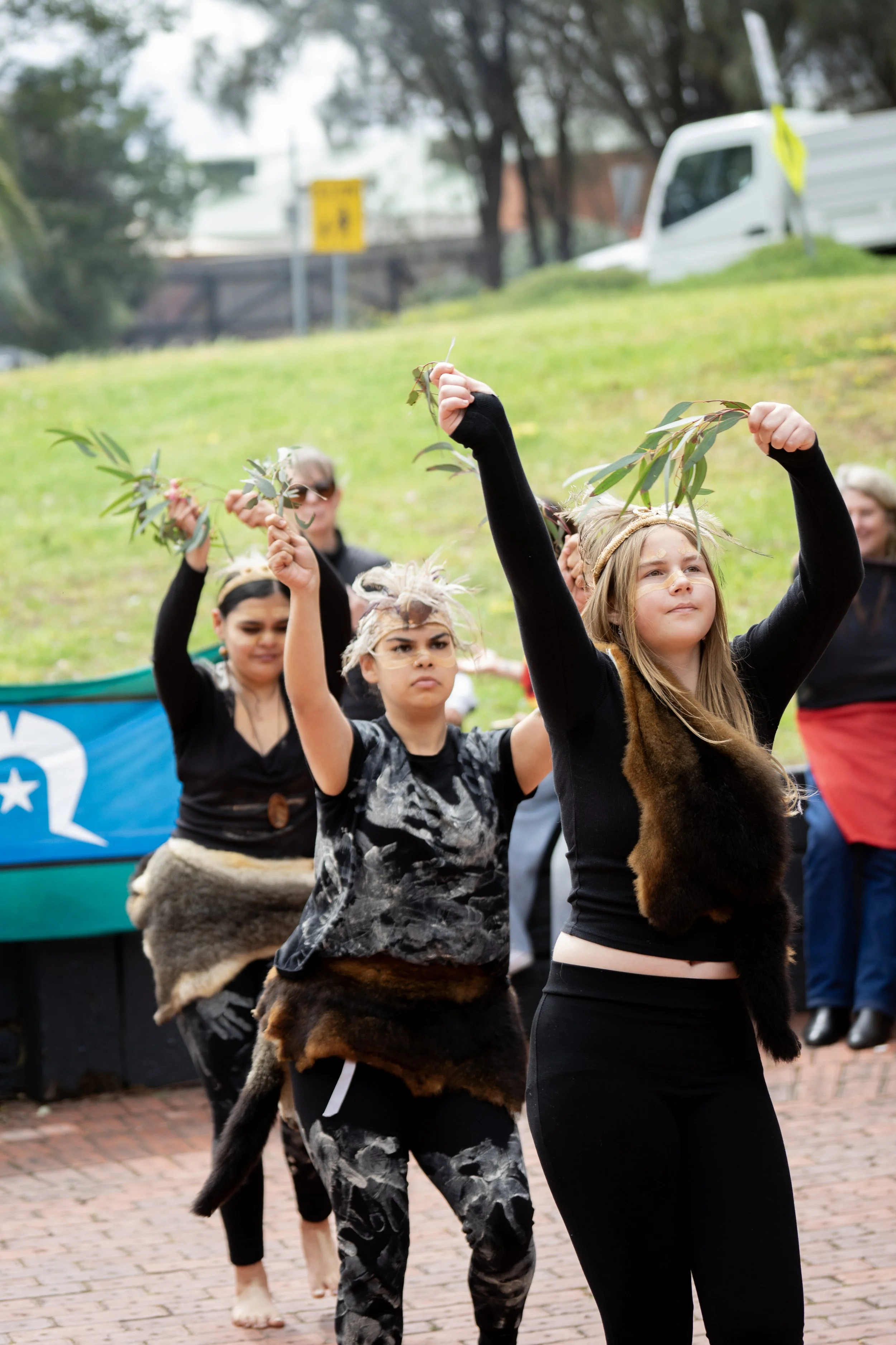 A group of young people dressed in traditional Indigenous clothing, participating in a cultural dance or ceremony outdoors, holding green leaves and raising their arms.