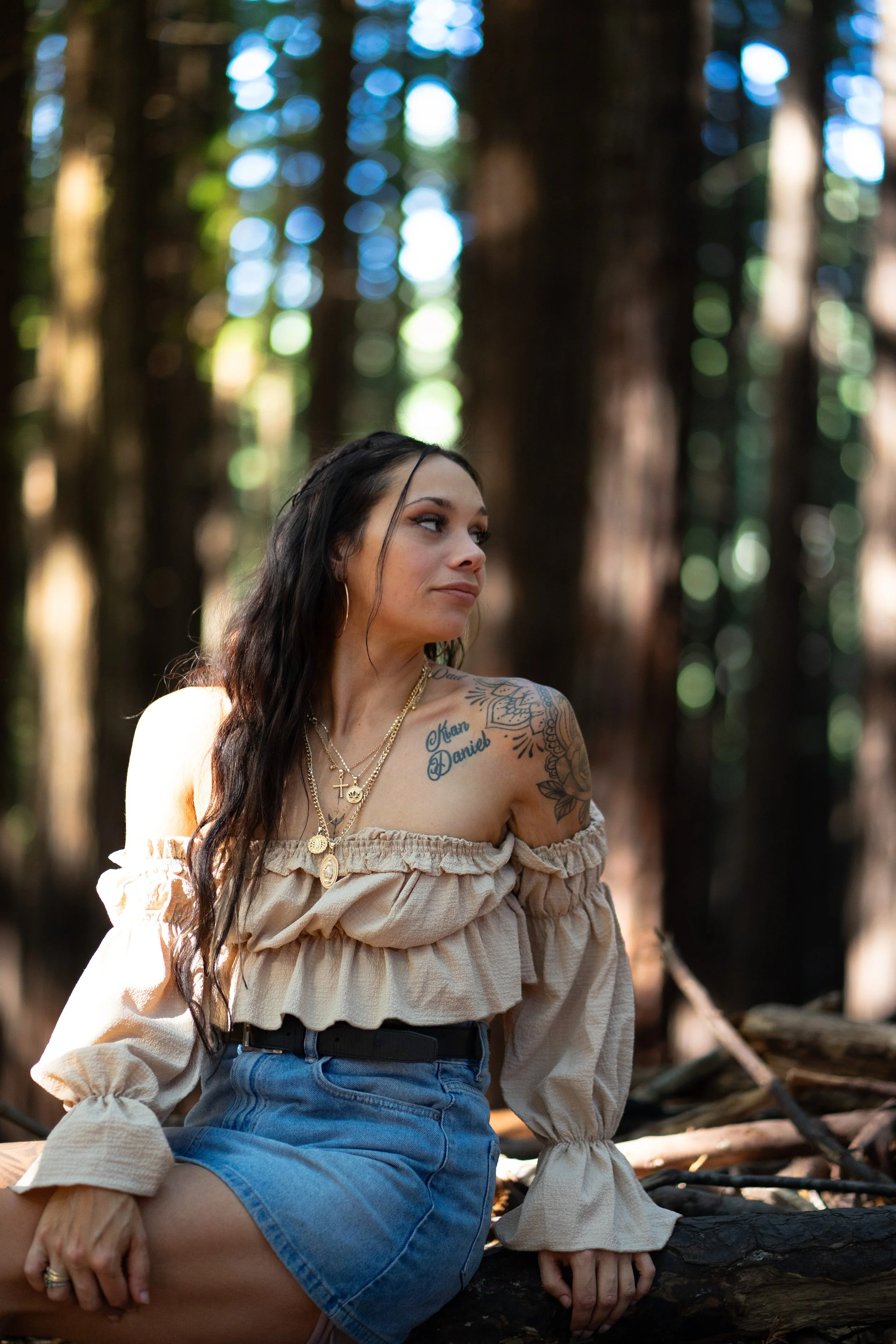A woman with long dark hair and tattoos on her shoulder, wearing an off-the-shoulder beige top and denim skirt, sitting outdoors in a forest with tall trees in the background.