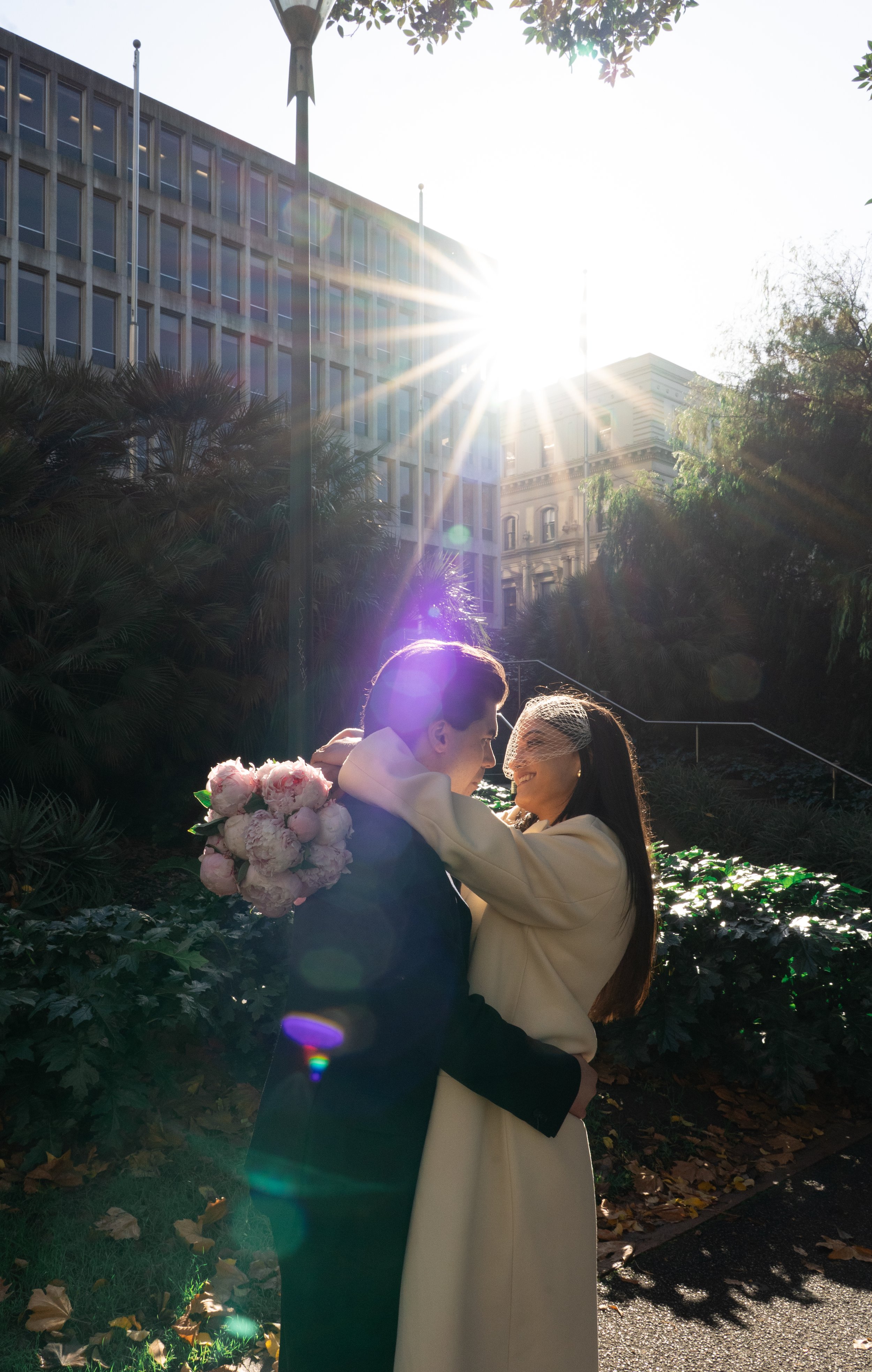 A couple dressed in formal attire, holding a bouquet of pink and white flowers, embracing outdoors with sunlight shining brightly behind them, creating a lens flare effect. The scene has trees, plants, and tall buildings in the background.