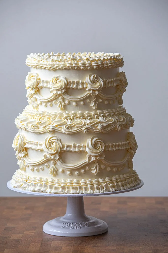 A tall, multi-layered white wedding cake decorated with piped flowers and ornate icing patterns, placed on a white cake stand.