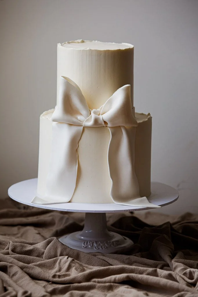 Two-tier white wedding cake with a bow decoration on the side, placed on a cake stand, with brown fabric underneath.