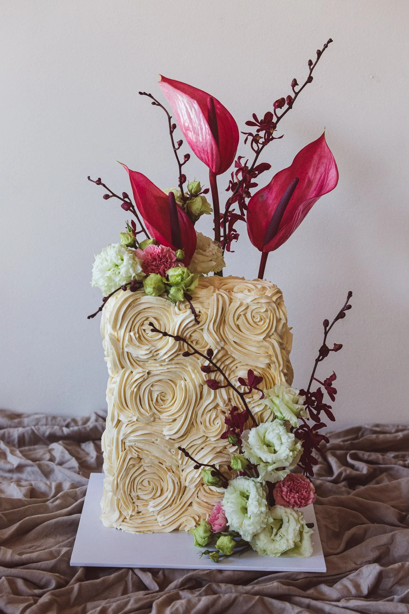 Elegant tiered cake decorated with cream-colored frosting in swirl patterns, adorned with pink anthuriums, white lisianthus, pink carnations, and dark red foliage, set on a white base with a beige fabric surface.