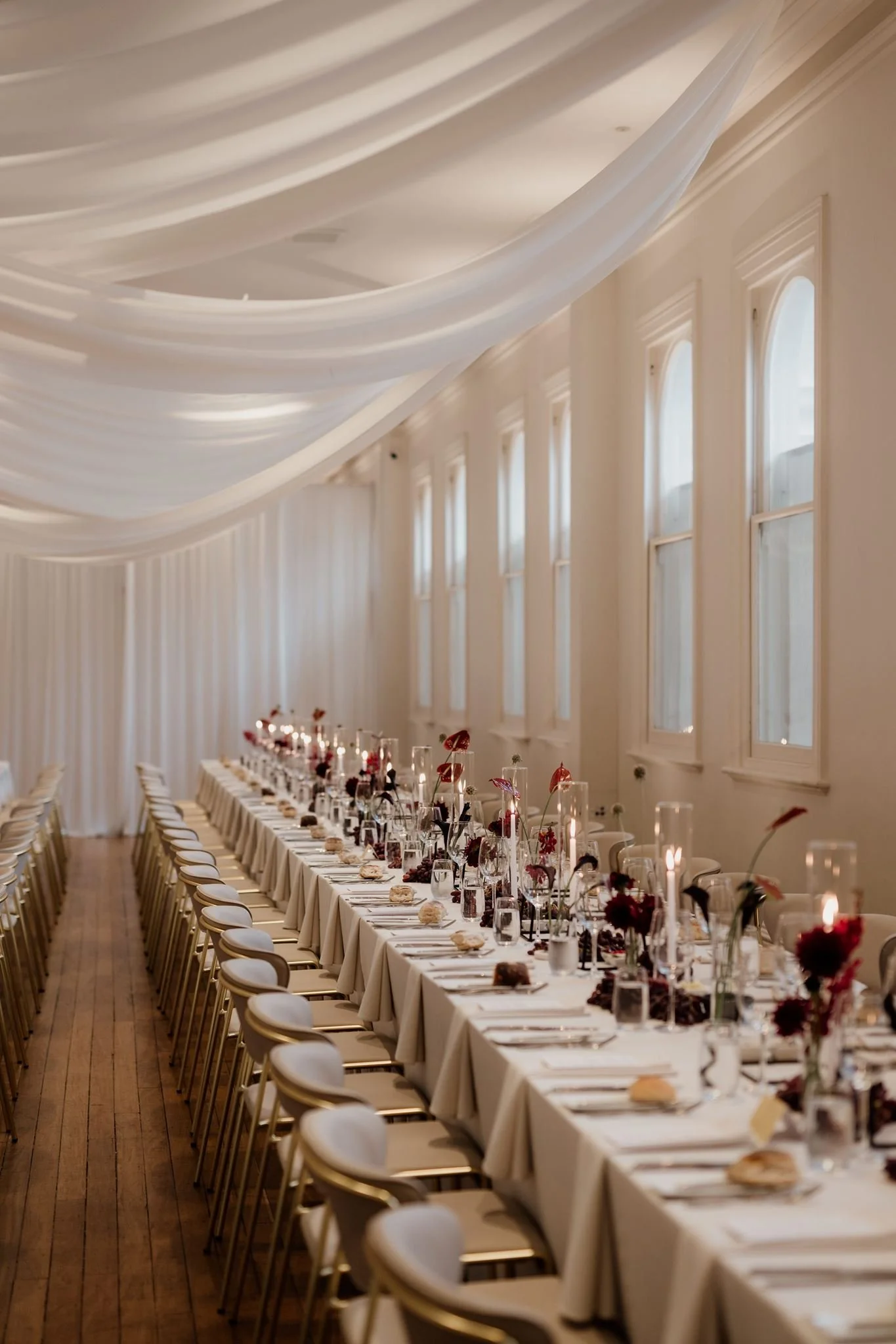 Long banquet table set with white tablecloths, floral centerpieces, candles, glassware, and tableware, in a high-ceiling room with white draped fabric and tall windows.