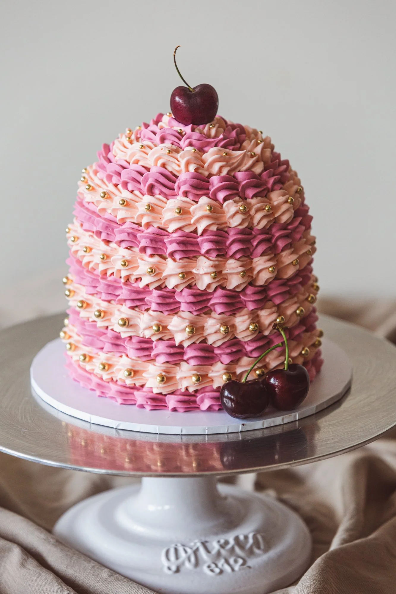 A tiered pink and purple frosted cake decorated with small golden balls and topped with cherries.