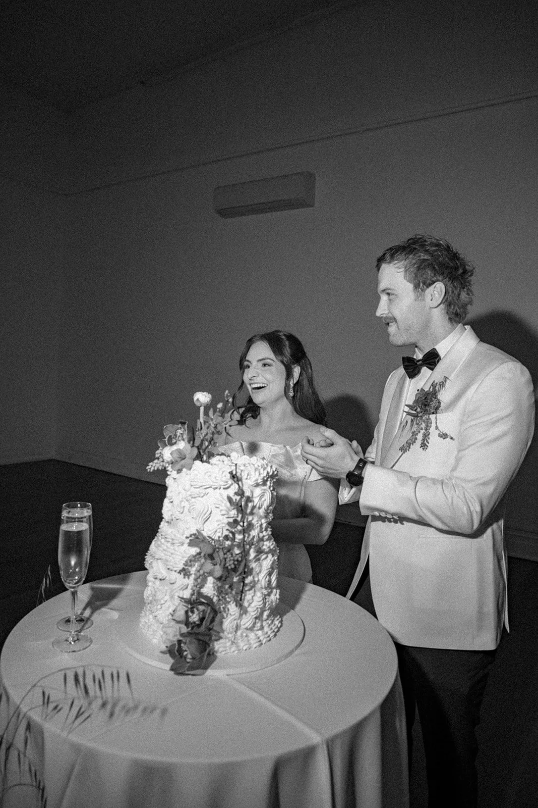 A bride and groom at their wedding reception, cutting a wedding cake. The bride is smiling and laughing, wearing an off-the-shoulder dress, and holding a small bouquet. The groom is wearing a light-colored tuxedo with a bow tie and boutonniere, clapp