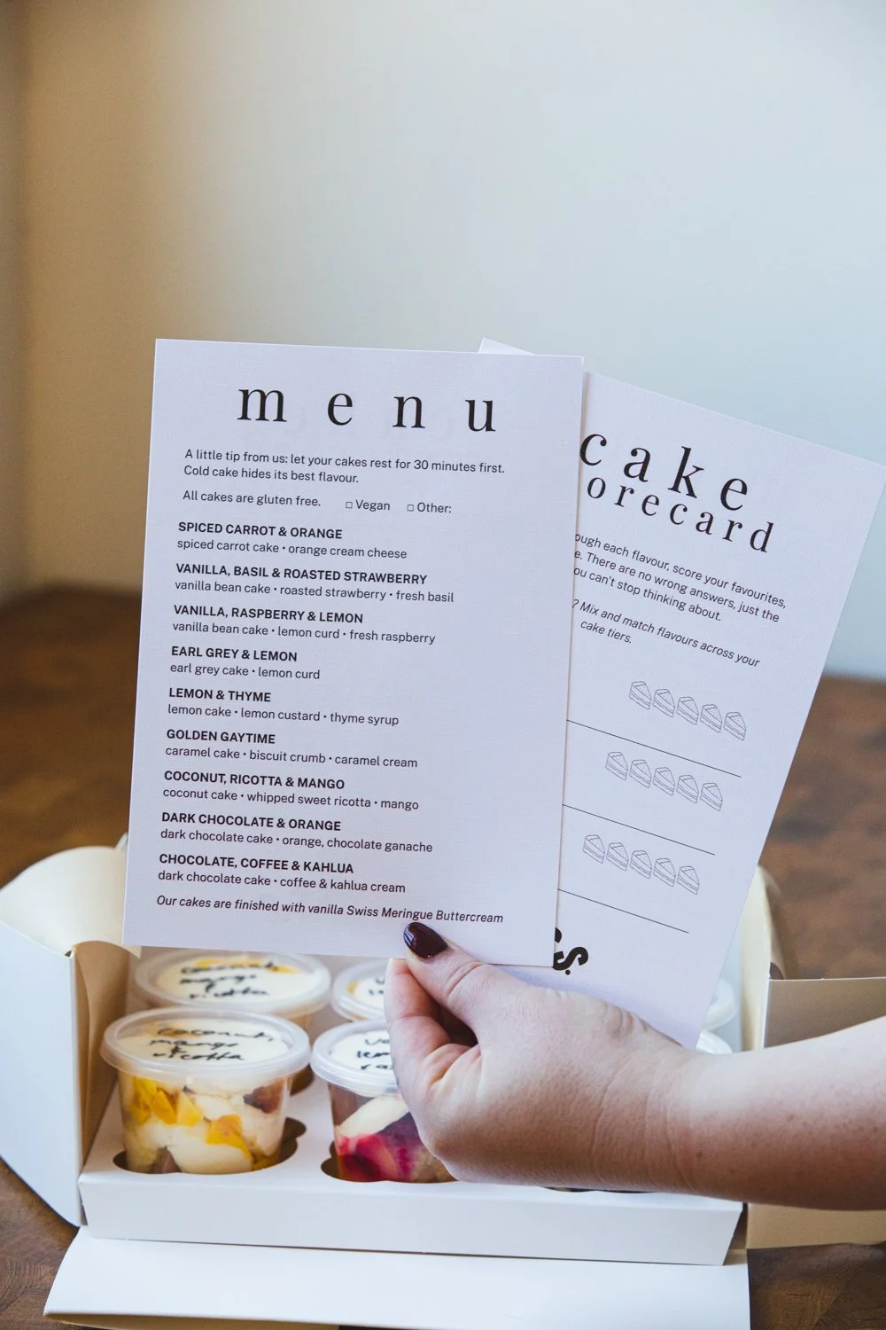 Person holding a cake menu and a cake card above a box of assorted cakes, including fruit and cream desserts.