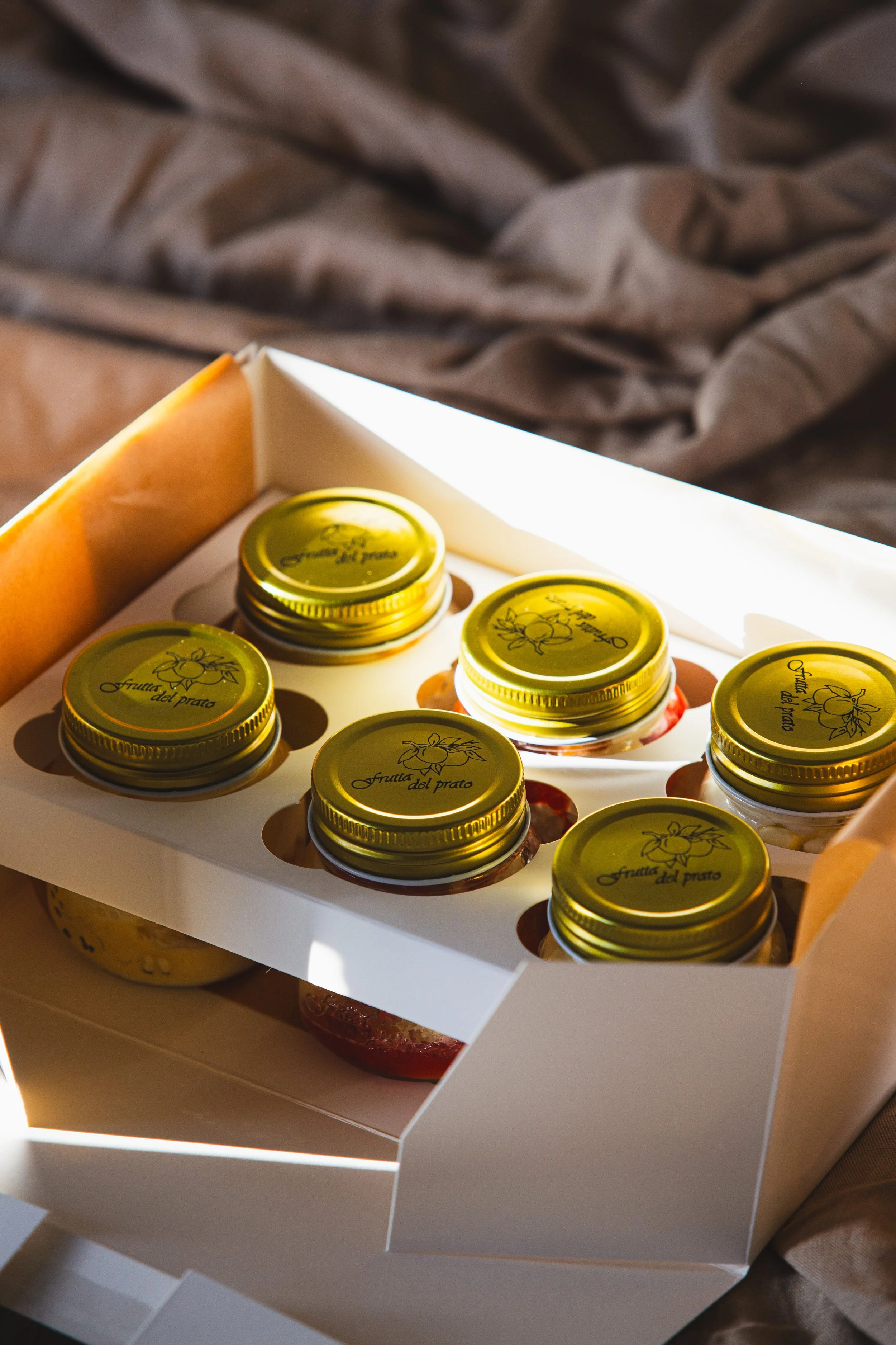 A box containing small jars with yellow lids, labeled "frutta del prato," sitting on a grey fabric surface.