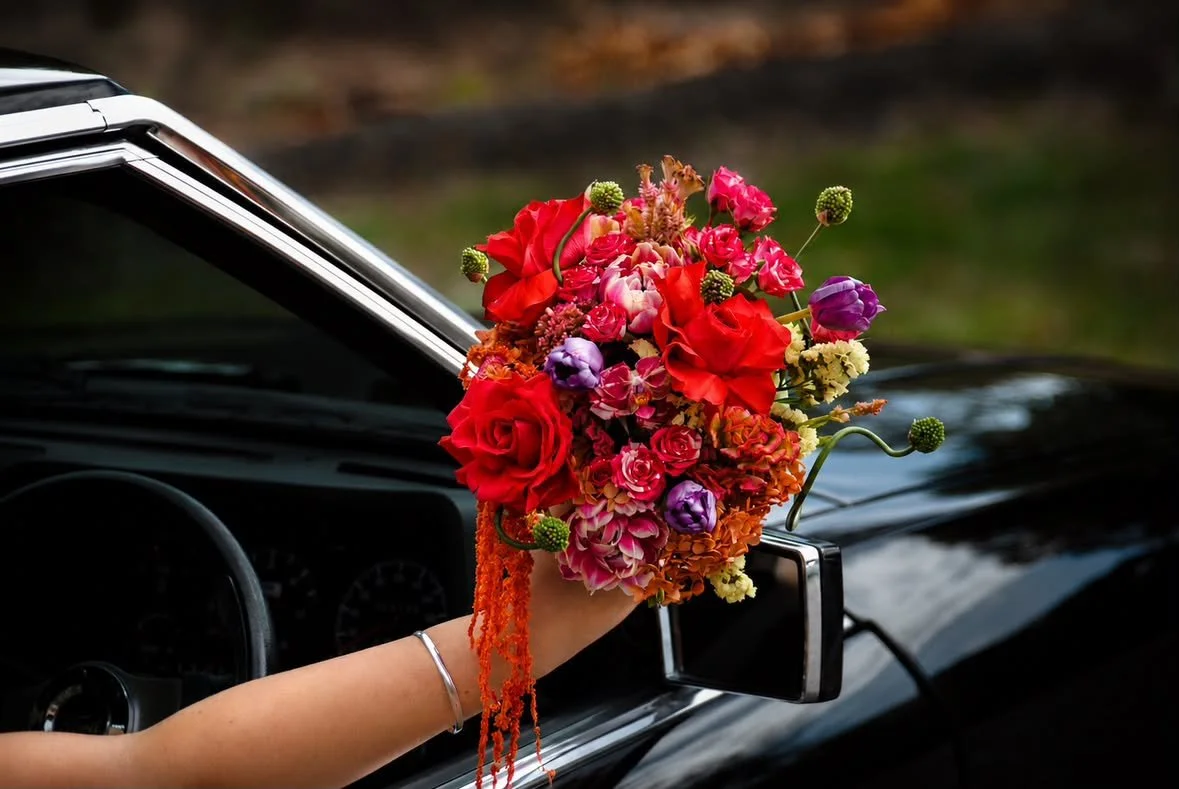 A person holding a colorful bouquet of mixed flowers next to the side mirror of a black car.