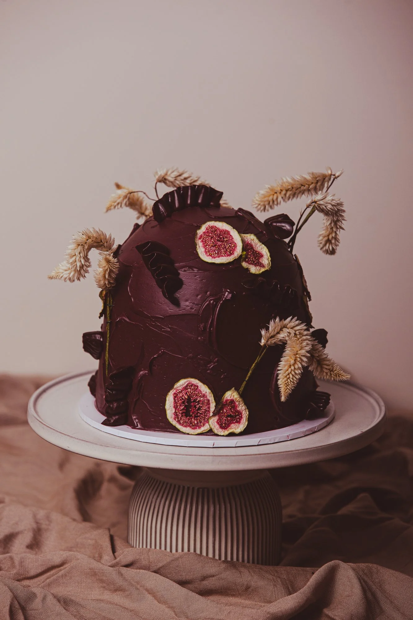 A dark purple, glossy, irregularly shaped cake with dried fig slices and beige dried flowers as decoration on a white cake stand, set against a neutral background.