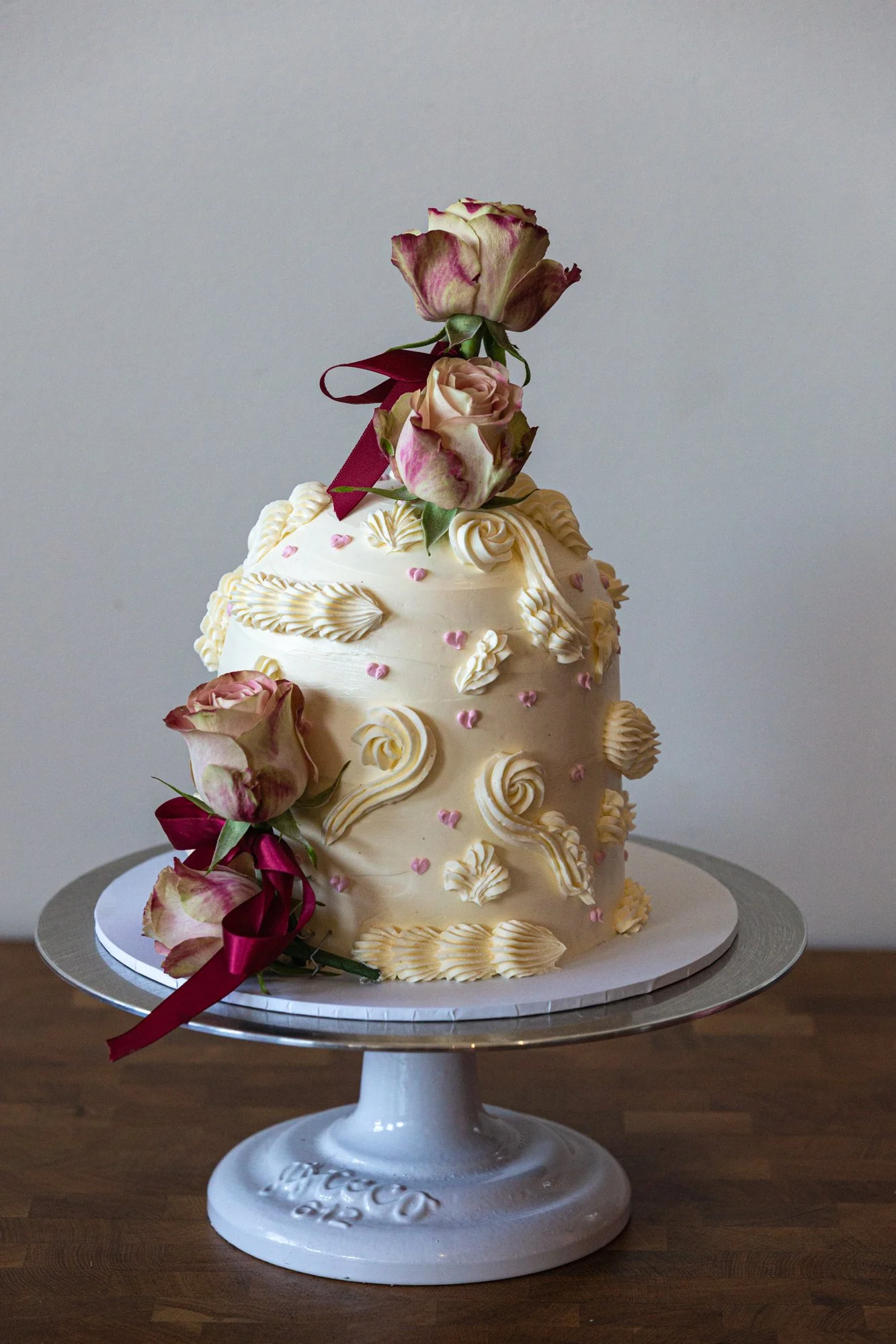 A decorated wedding cake with white icing, pink roses, and cream-colored piping, topped with roses and tied with maroon ribbons on a silver cake stand.