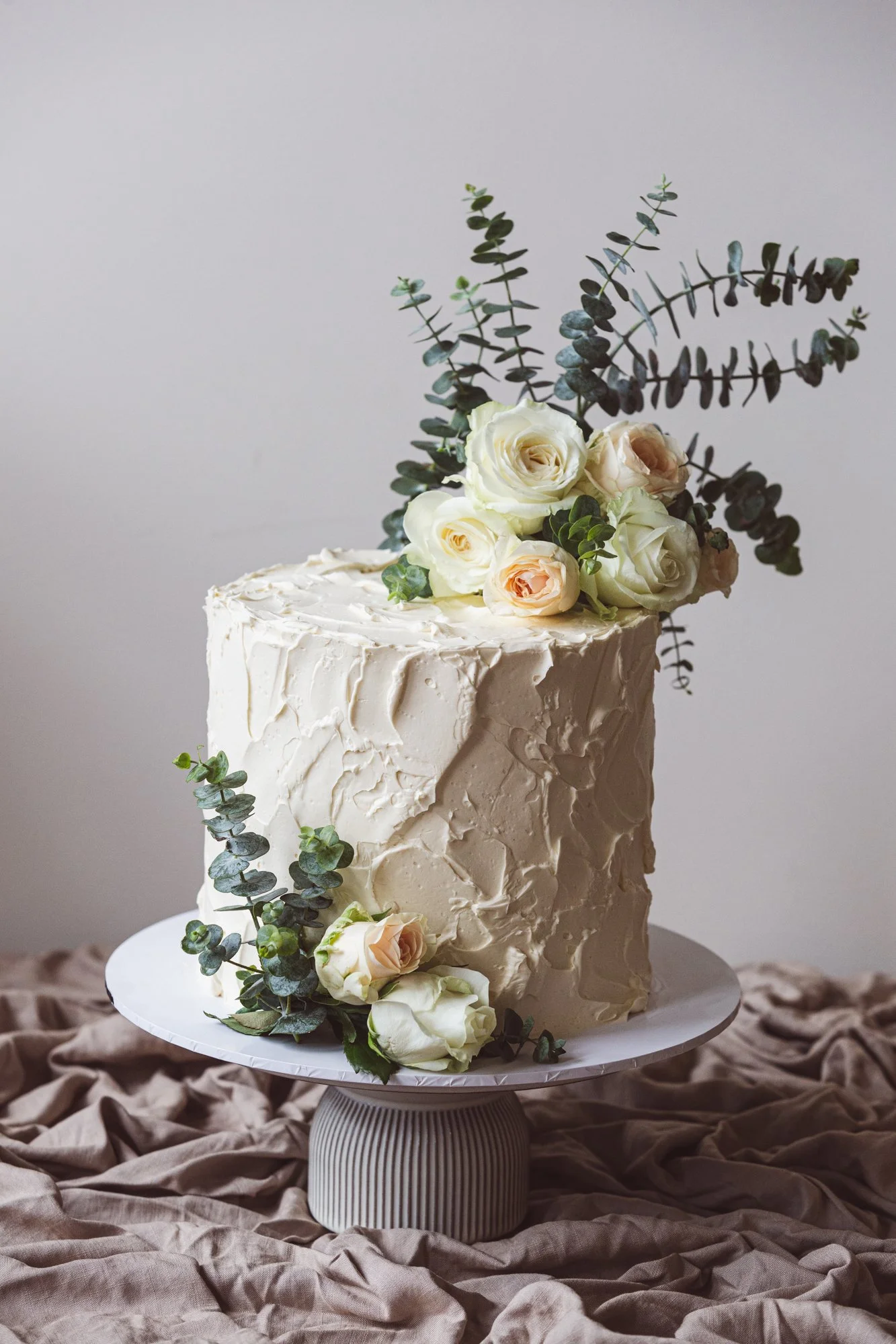 A tall, rectangular wedding cake with cream-colored frosting, decorated with white and blush roses, and eucalyptus leaves, placed on a white cake stand on a draped beige fabric background.