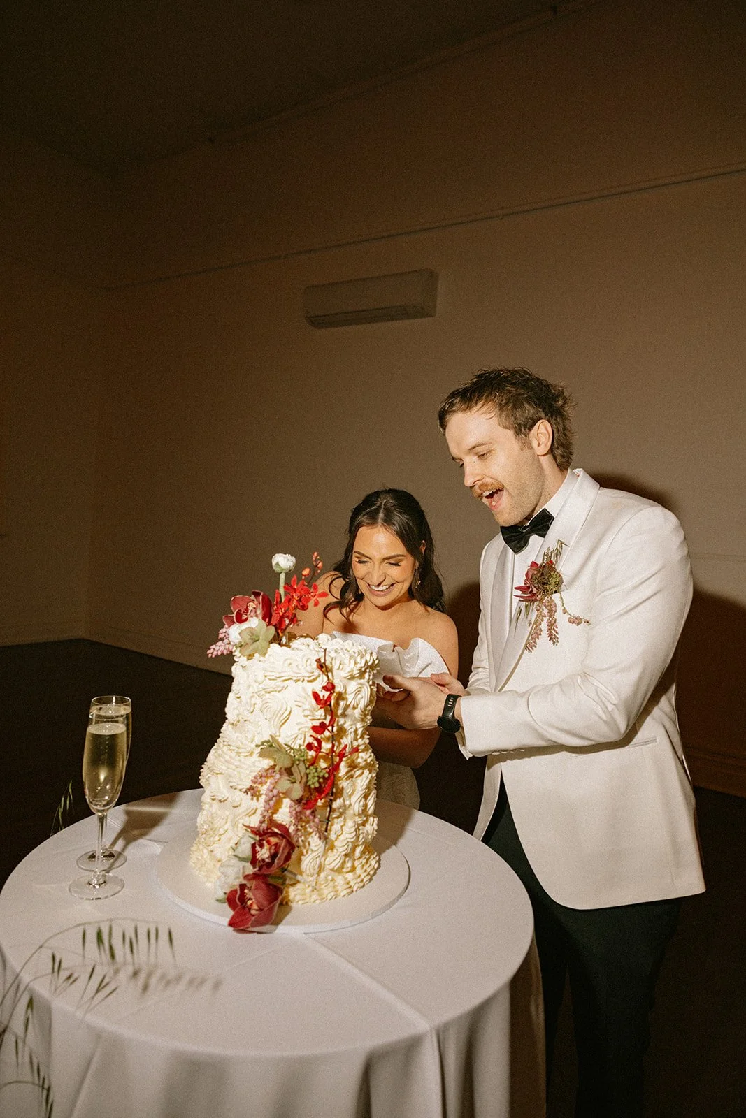 A bride and groom cutting a wedding cake together, smiling, with a champagne glass on a table.