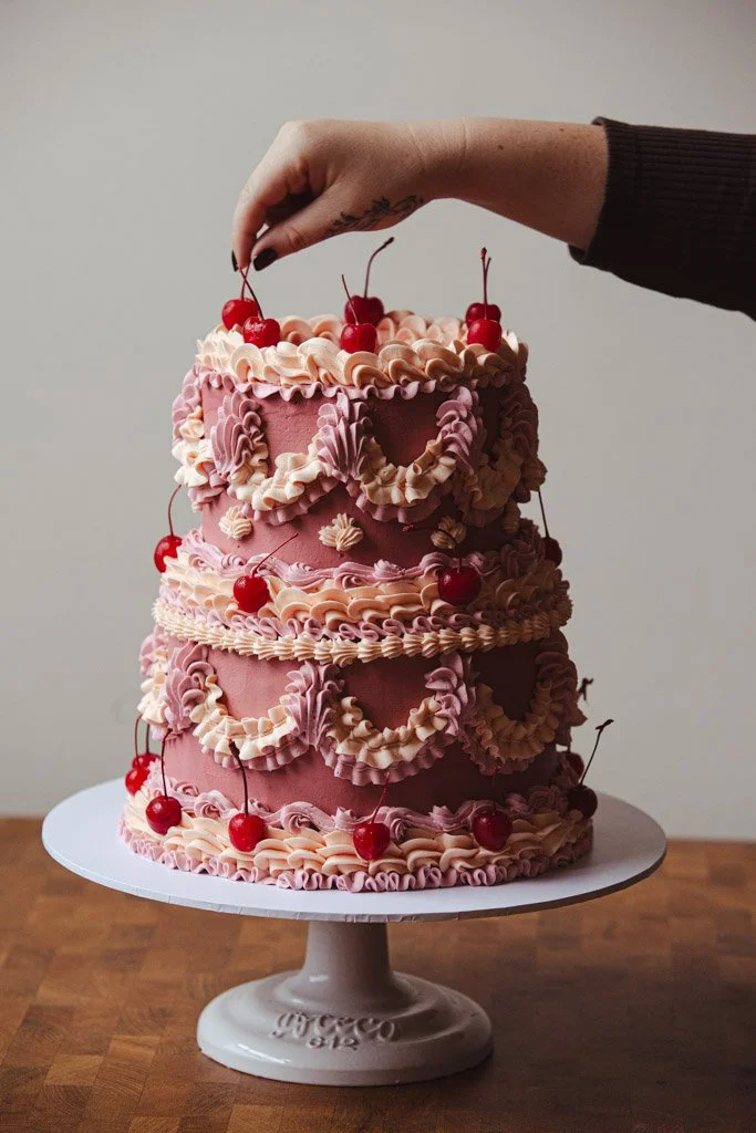 Three-tier pink cake decorated with pink and white piped frosting and topped with cherries, placed on a white cake stand on a wooden table.