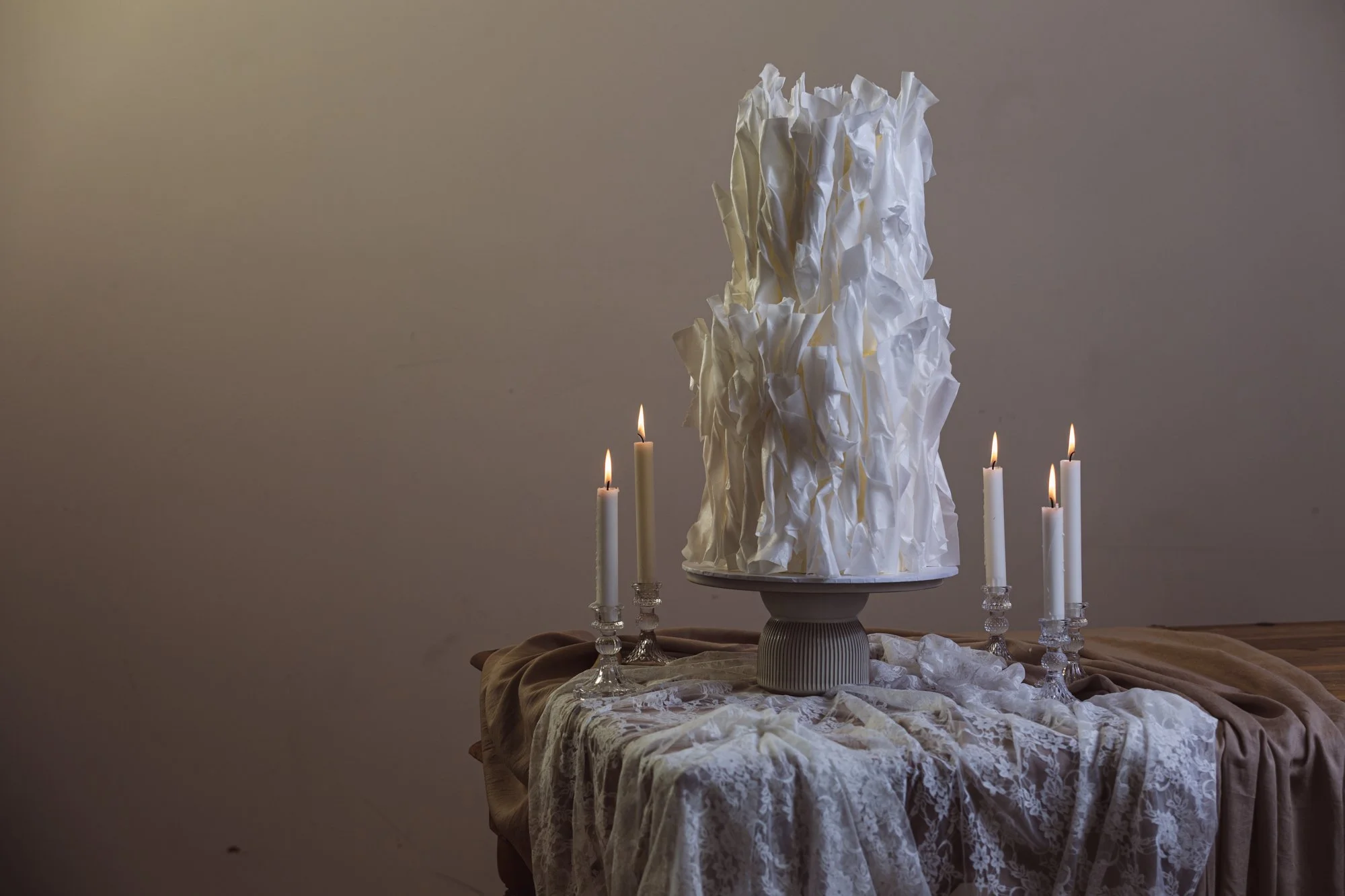 Table with crumpled white paper sculpture, surrounded by lit white candles, draped with beige and lace cloth, against a plain wall.