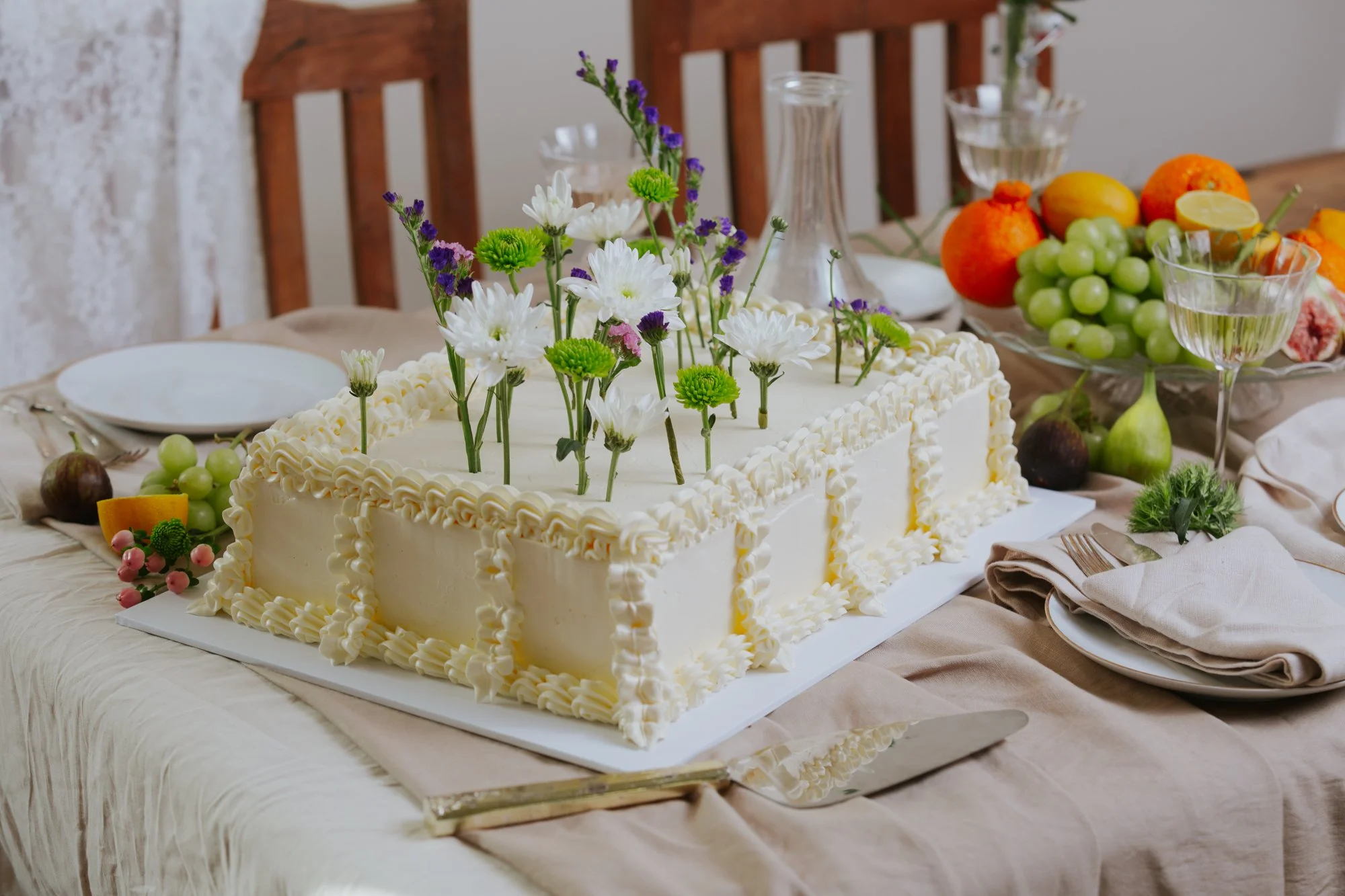 A rectangular white birthday cake decorated with white and purple flowers on top, placed on a table with various fruits and glassware around it.