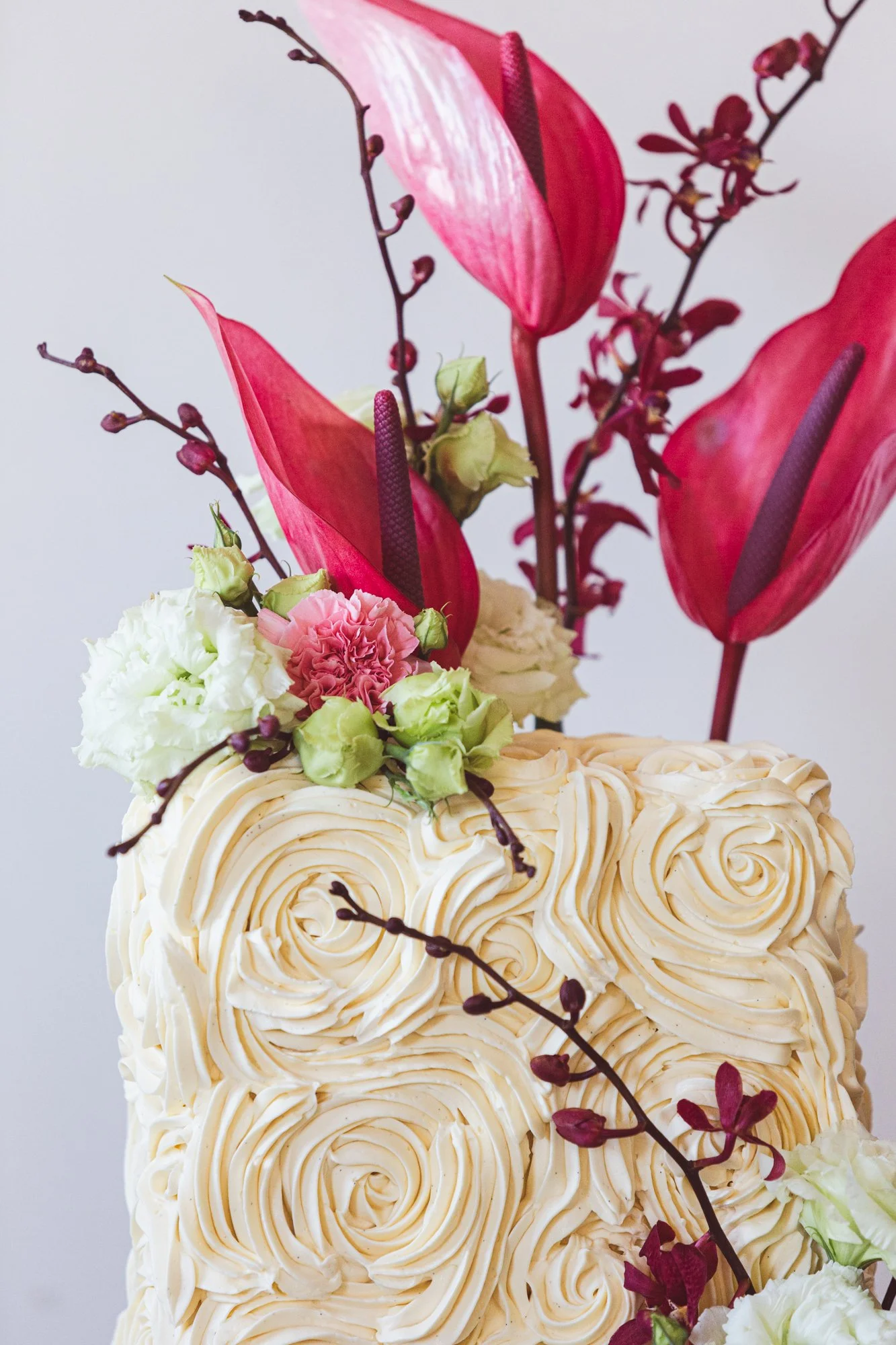 Close-up of a cake decorated with cream swirls and topped with pink and white flowers and dark purple anthuriums, with branches of tiny dark purple buds.