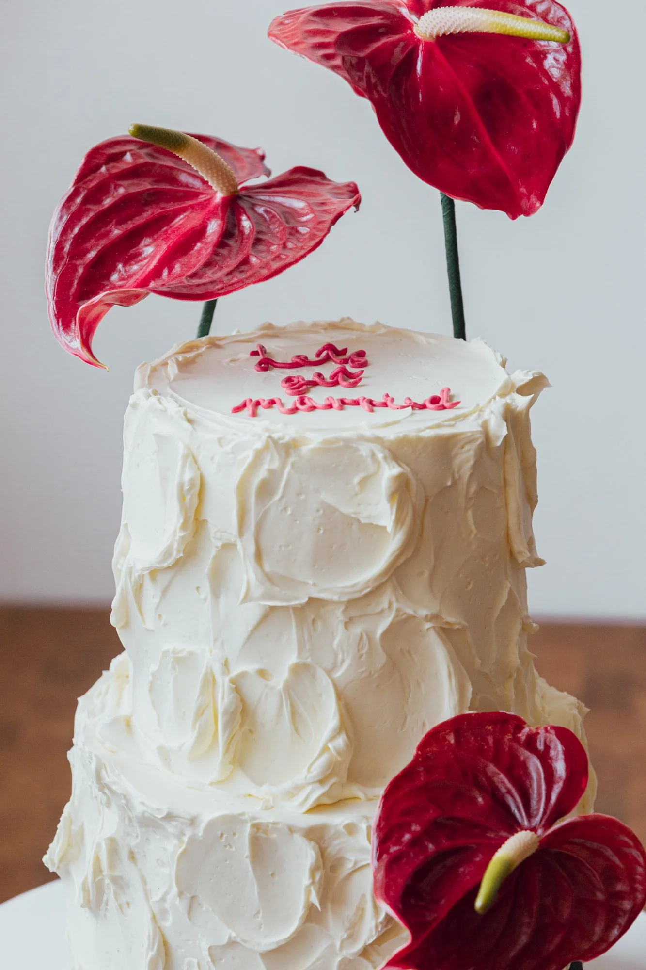 A multi-tiered white frosted cake decorated with red anthurium flowers and a small pink inscription on top.