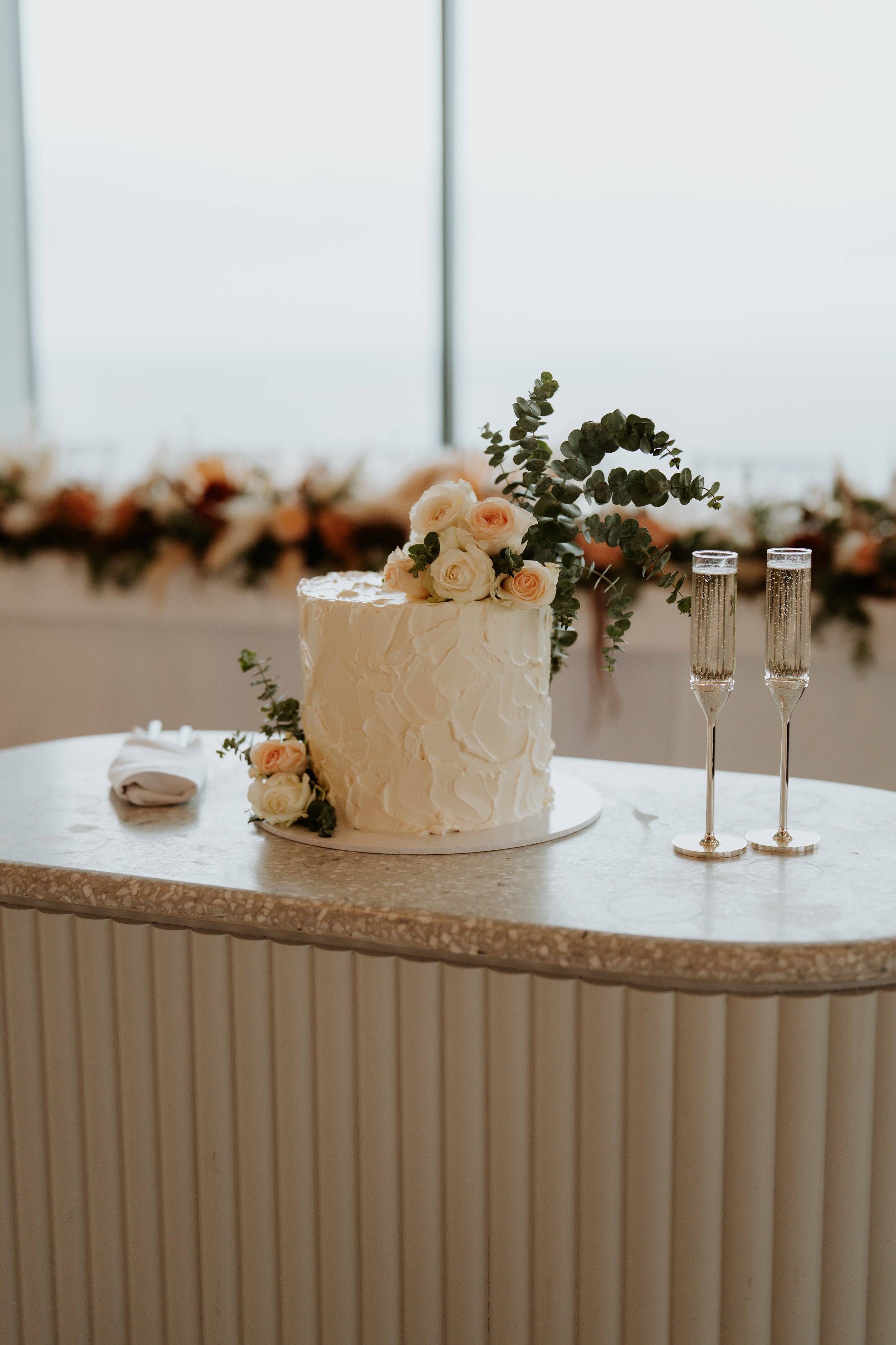 A white wedding cake decorated with pale pink and white roses and eucalyptus leaves, two champagne glasses filled with bubbly, all placed on a speckled marble countertop with vertical striped paneling beneath, and a blurred background with floral decor.