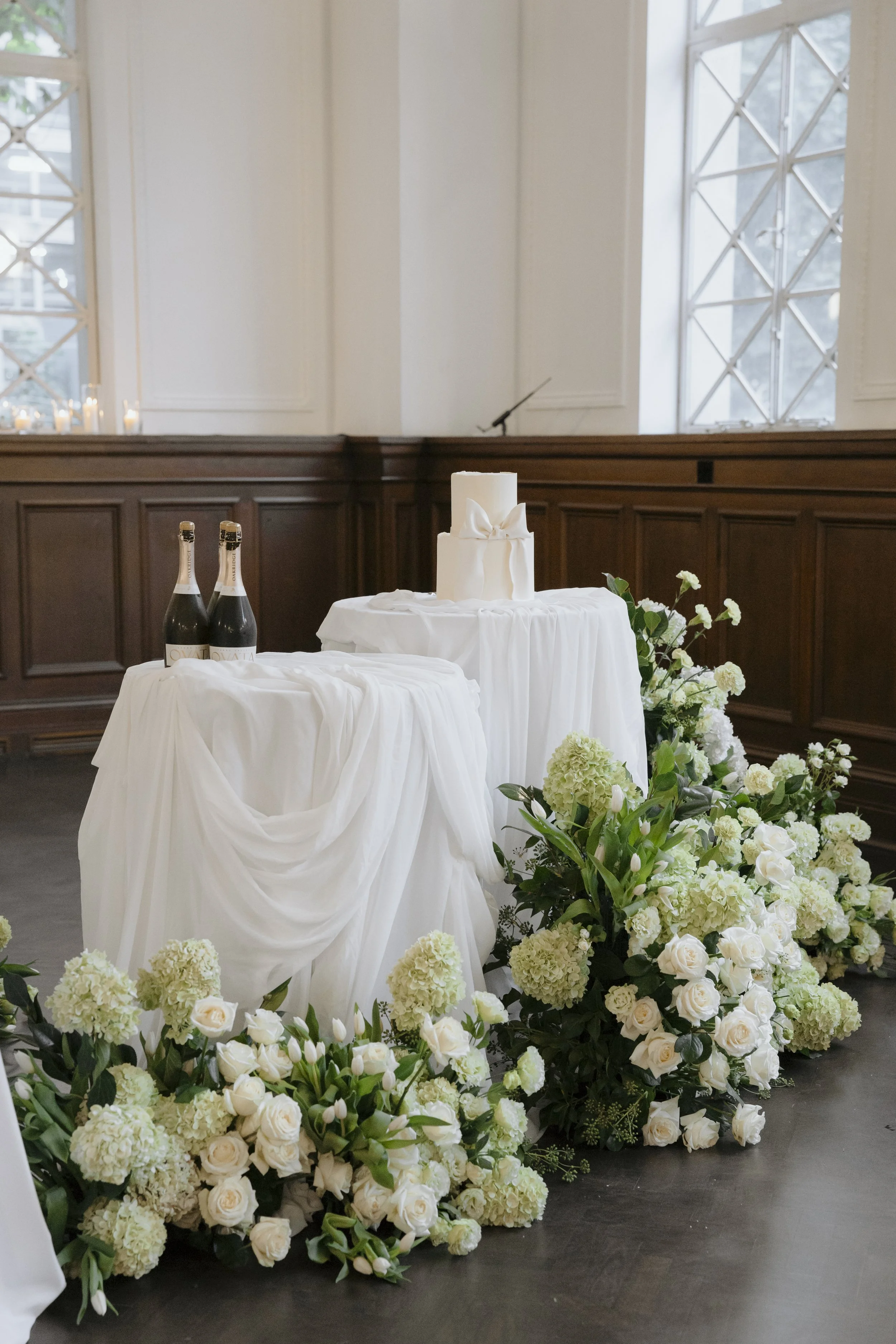 A wedding altar setup with a white cloth-draped table holding a white candle with a bow, surrounded by floral arrangements of white roses, hydrangeas, and greenery, in a room with wooden wainscoting and large windows.
