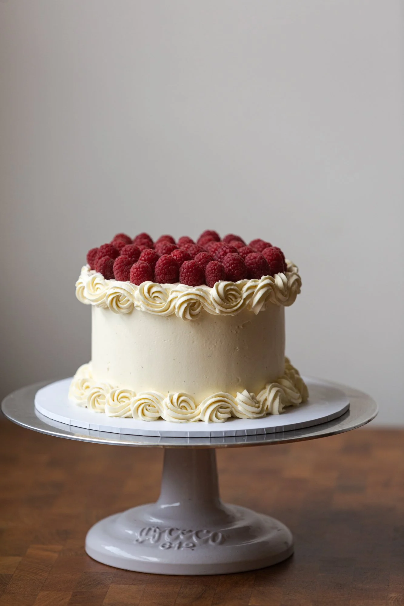 Layered white cake decorated with whipped cream and fresh raspberries on top, on a white cake stand.