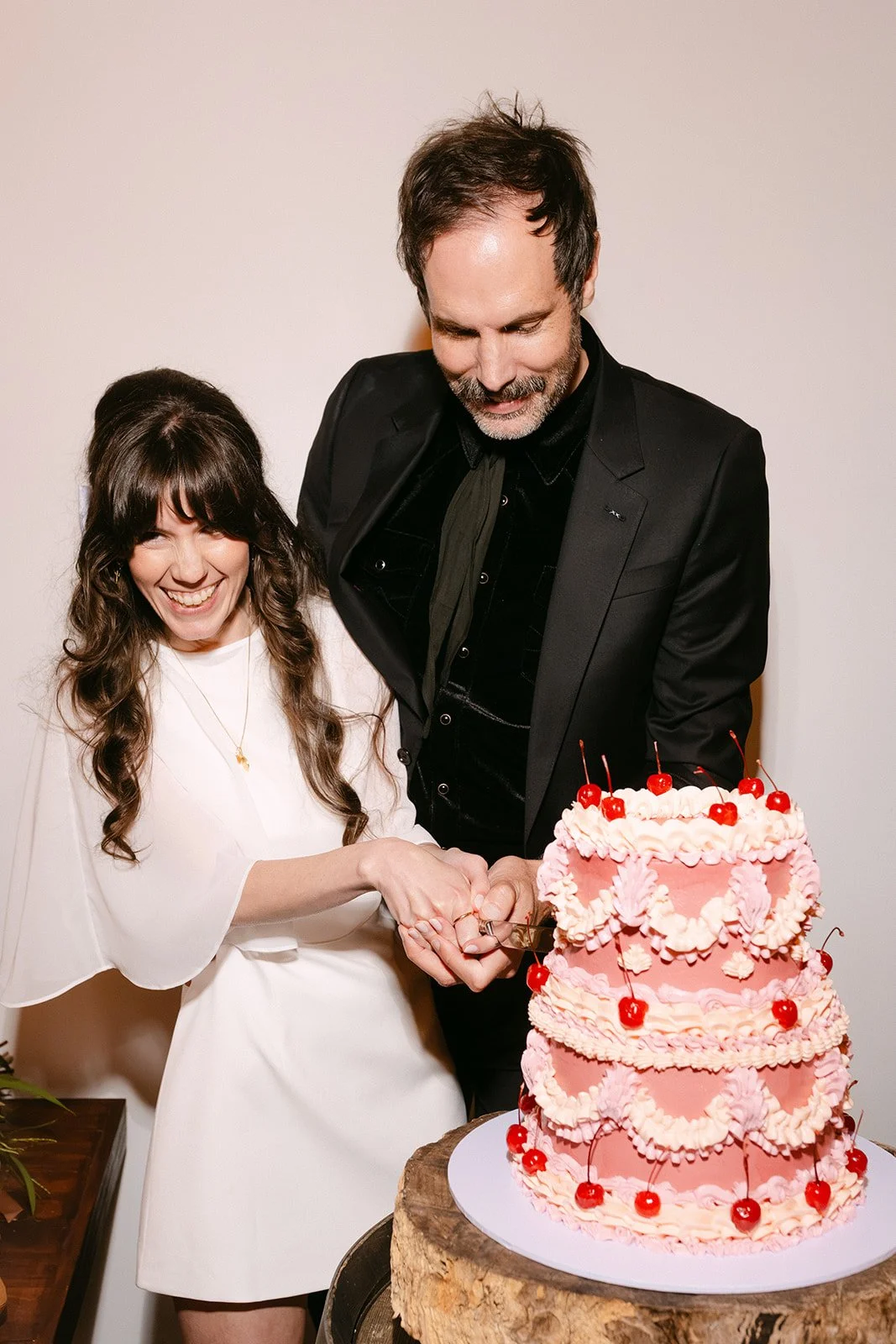 A woman and a man cutting a large, pink, three-tiered cake with cherries on top, at a celebration.