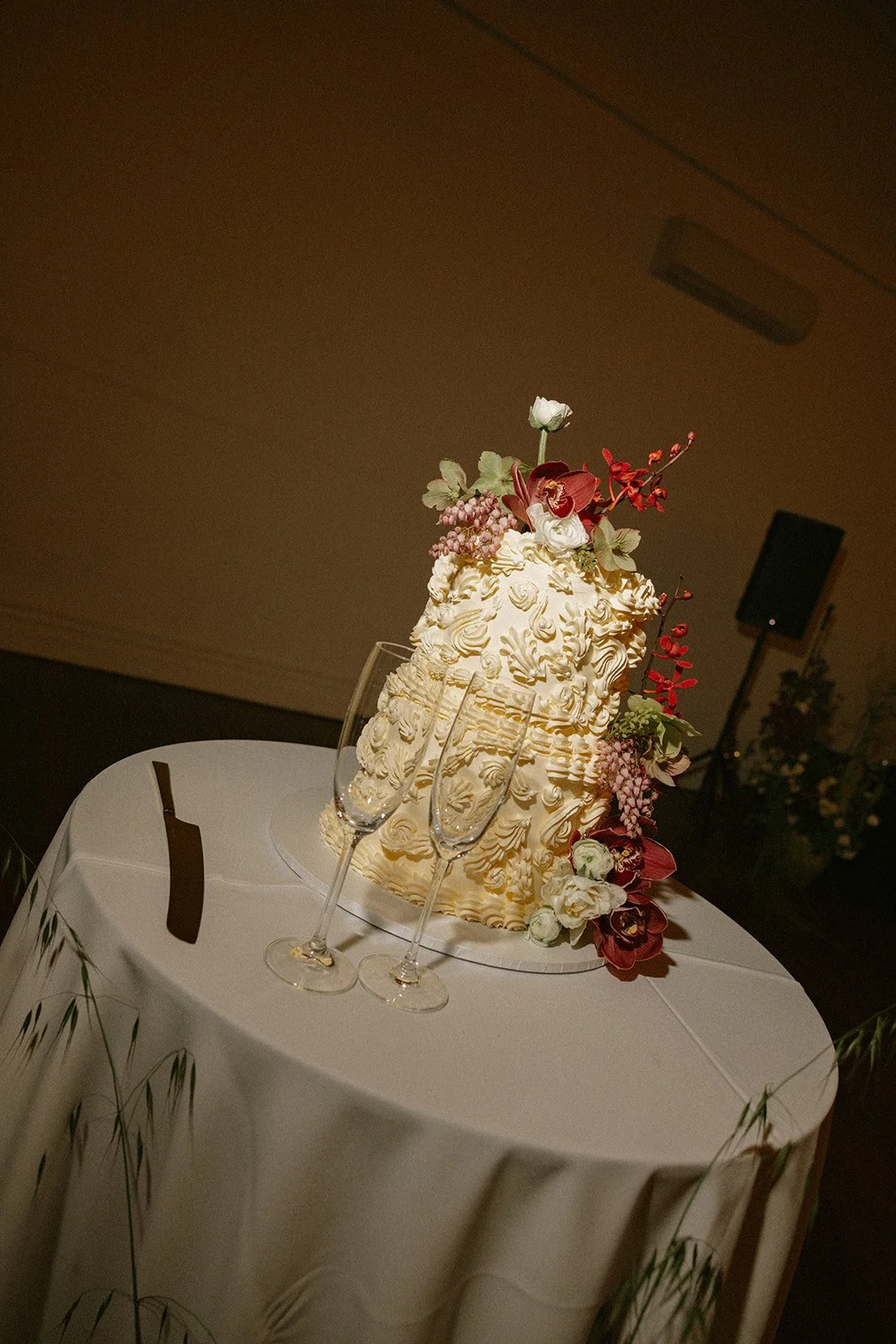 A decorated wedding cake with white and floral decor, three champagne flutes, and a black card on a round table covered with a white tablecloth at an indoor event.