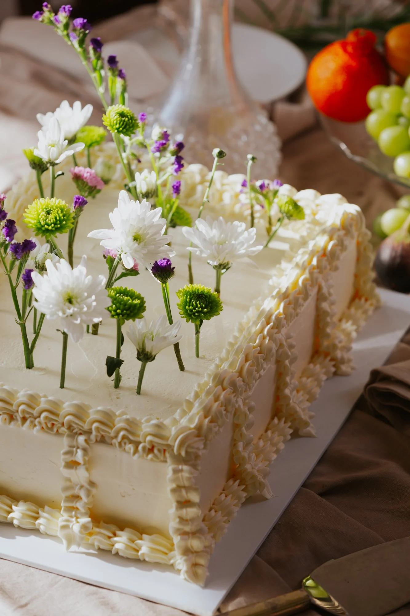 A wedding cake decorated with fresh white, purple, and green flowers, with a heap of cream border details around the edges. In the background are fruits including watermelon and grapes.