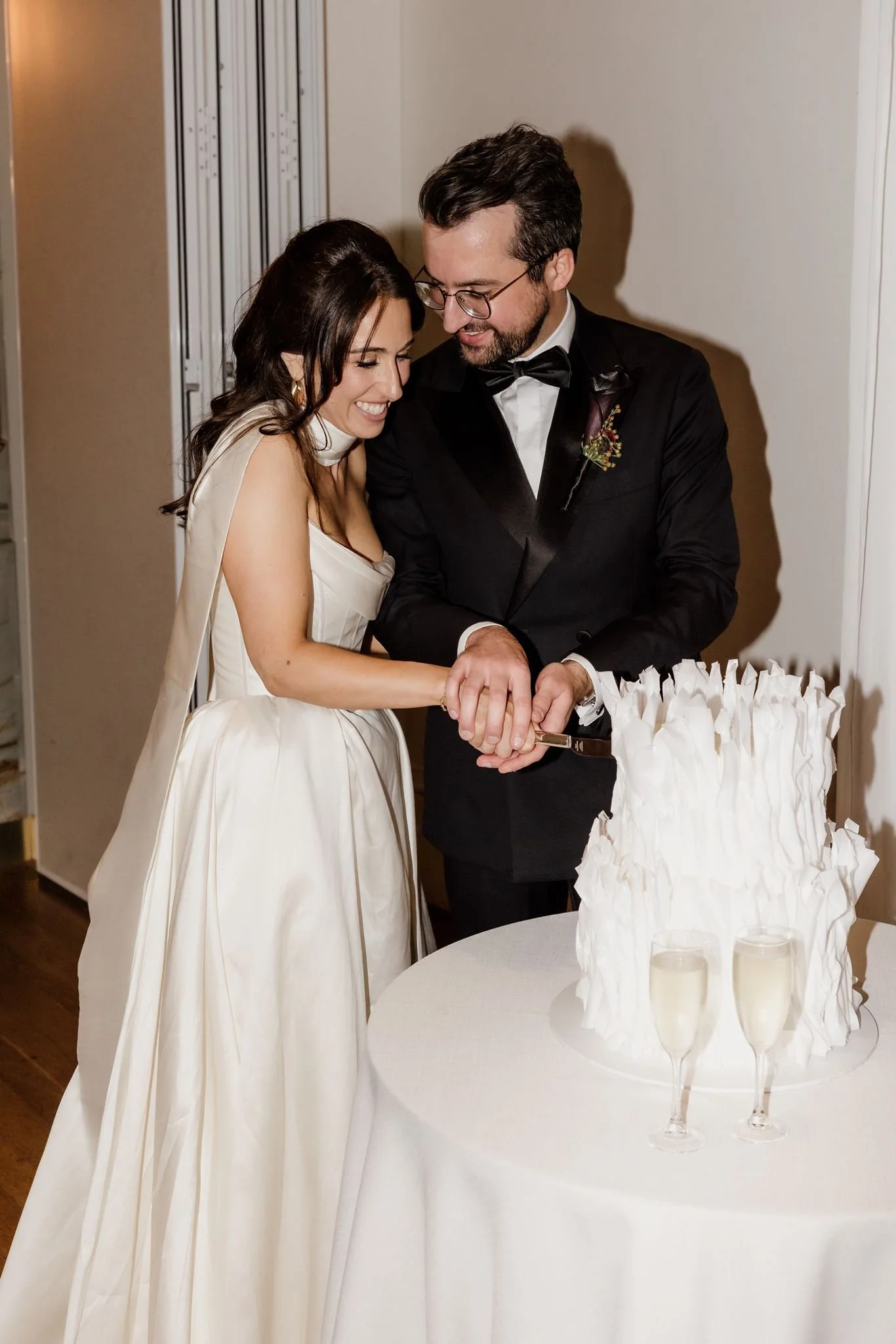 A bride and groom smiling and holding a knife together to cut a wedding cake, standing next to a round table with two glasses of champagne.