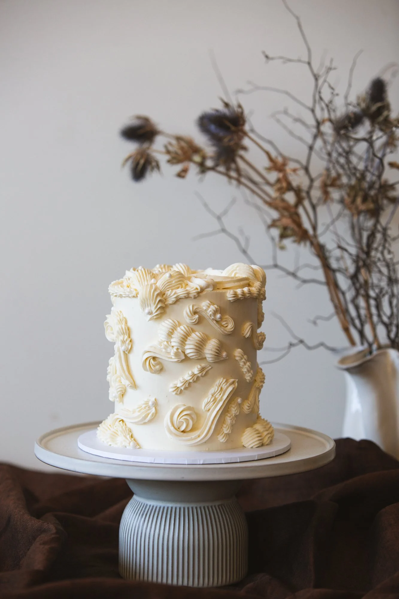 A tall, elegant white cake decorated with intricate buttercream piping, placed on a round cake stand, with a dried flower arrangement in a white vase in the background.