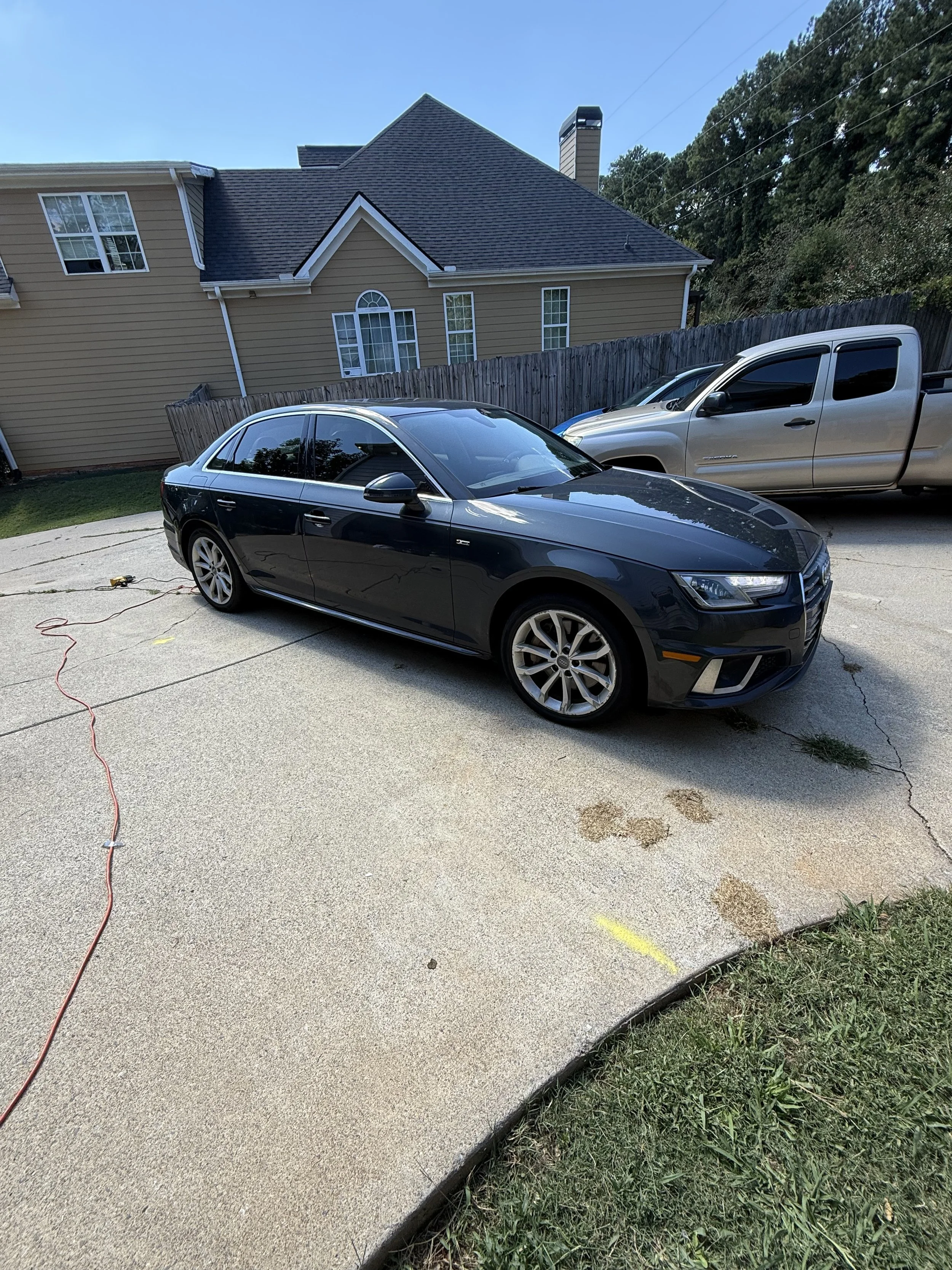 A black sedan parked on a concrete driveway next to a tan house, with a silver pickup truck nearby and a wooden fence in the background.