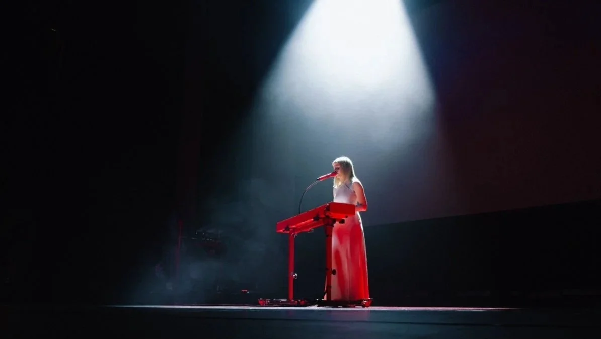 A woman in a long white dress performing on stage under a spotlight, playing a keyboard and singing into a microphone.