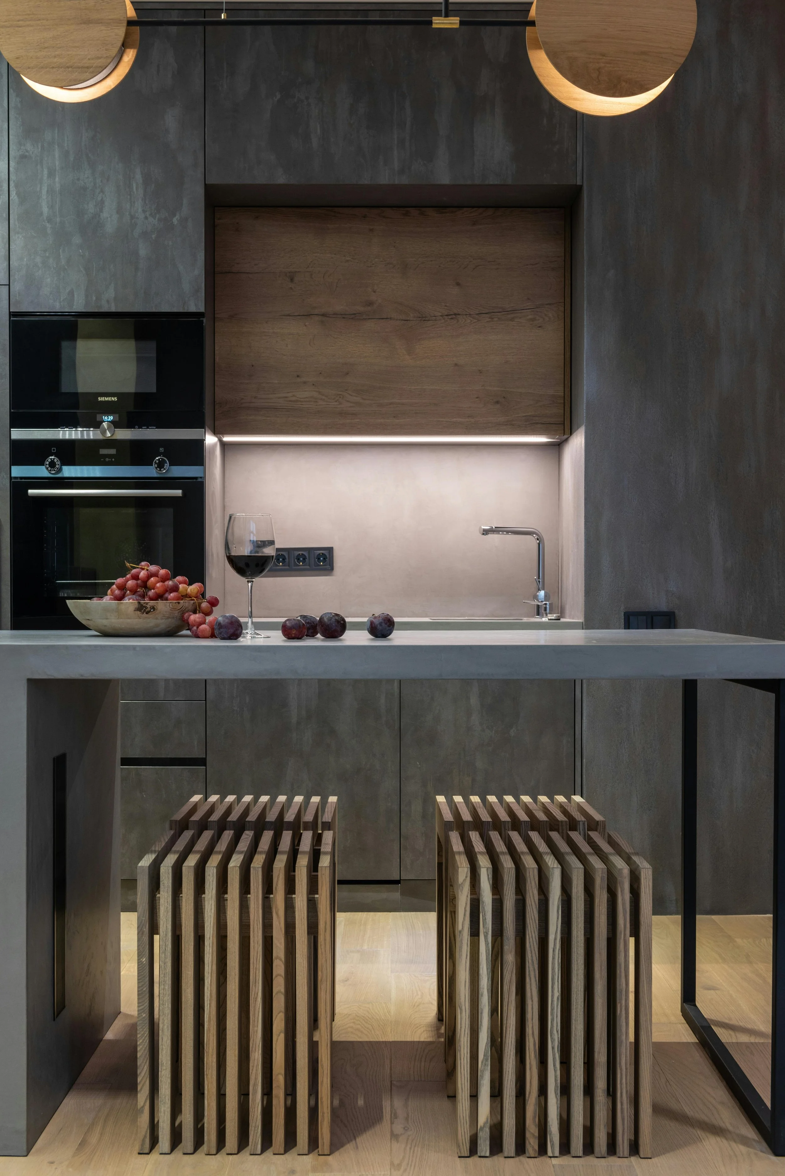 Modern kitchen interior with minimalist design, featuring a concrete countertop, wooden stools, grapes, plums, and a wine glass on the counter. Dark cabinetry and built-in oven in the background, under-cabinet lighting, and a faucet at the sink.
