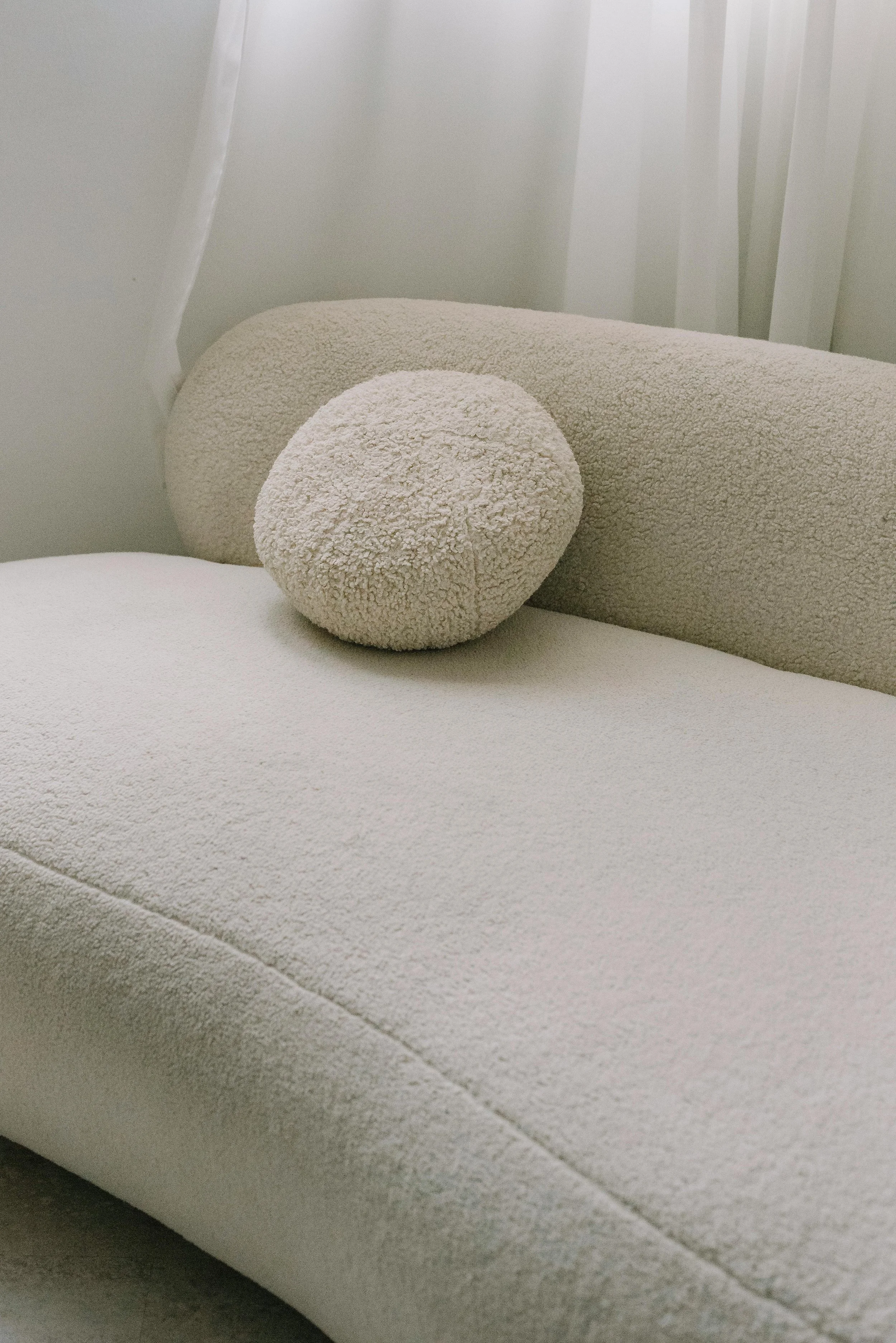 Beige boucle sofa with round pillow near sheer curtains.