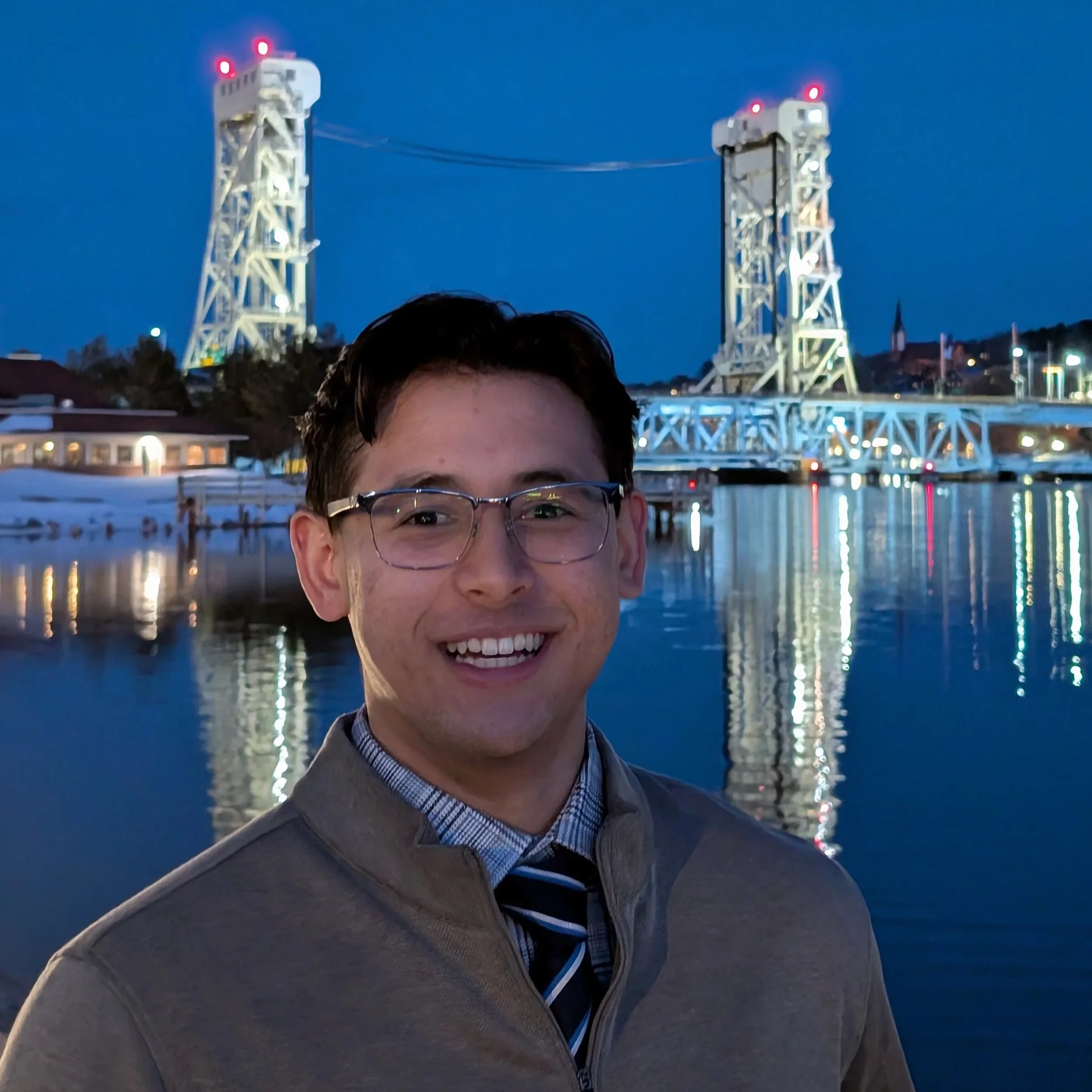A smiling man wearing glasses, a brown jacket, plaid shirt, and tie standing in front of a body of water at dusk with a bridge illuminated in the background.
