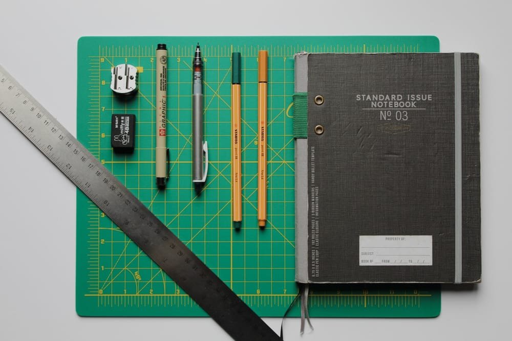 A green cutting mat with various stationery items including a ruler, eraser, two pens, two pencils, a mechanical pencil, and a black notebook with a gray elastic band, all arranged on it.