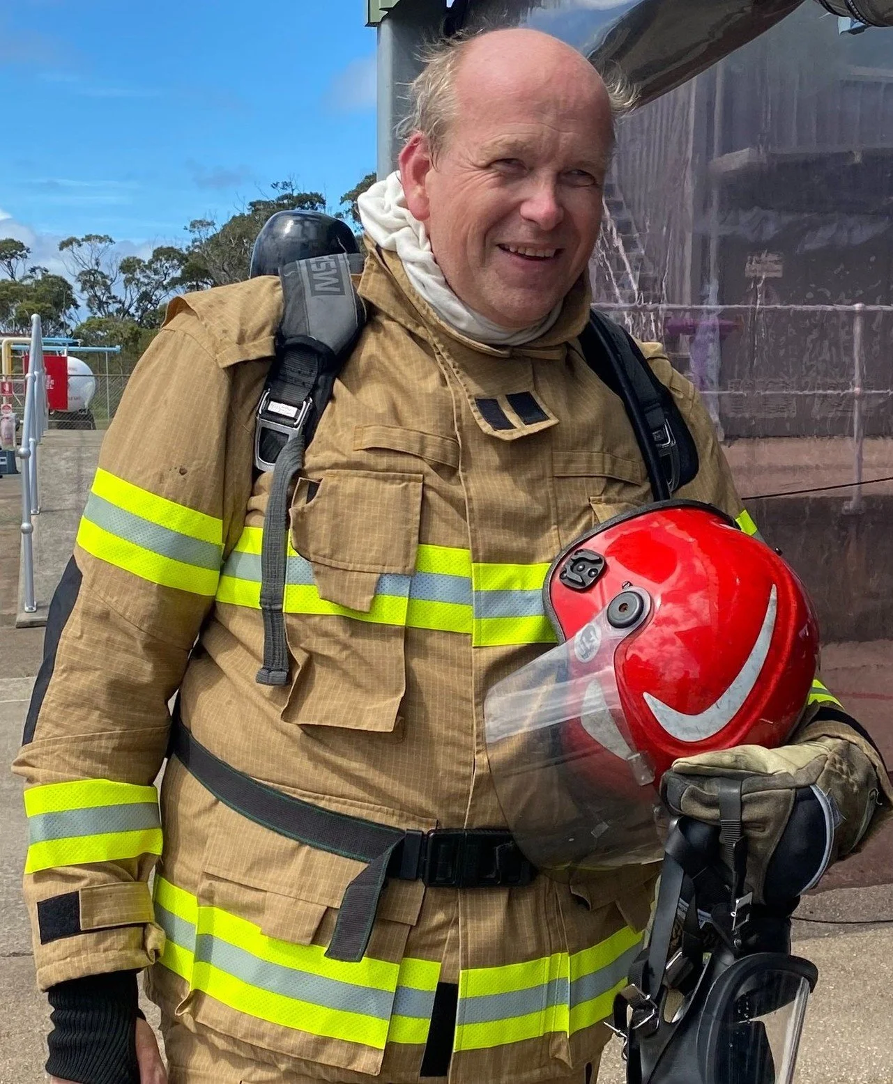 A smiling firefighter in full gear holding a red helmet with a gray visor and Nike logo, standing outdoors during daytime.