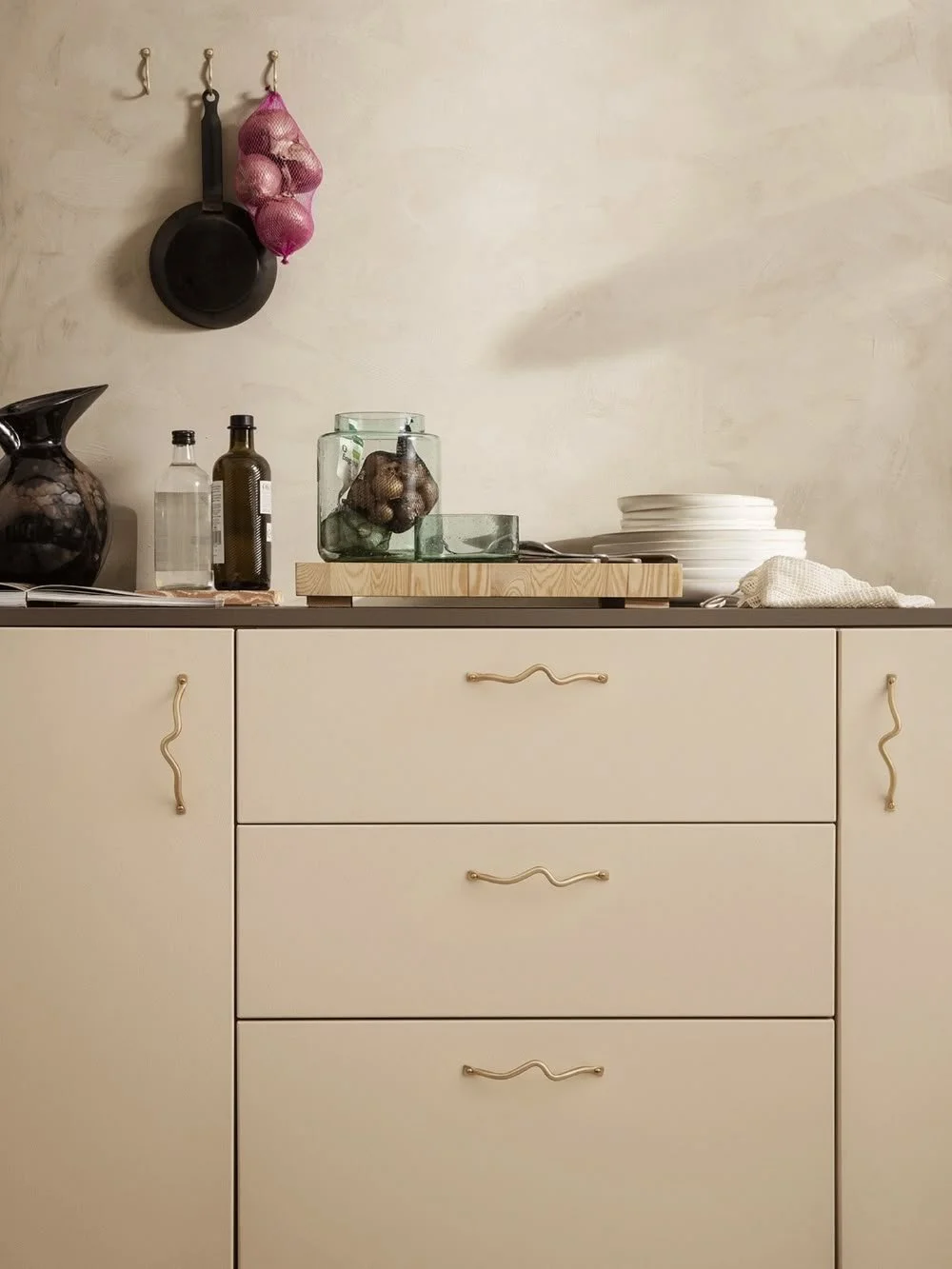 Kitchen countertop with beige cabinets, wavy handles, hanging frying pan, mesh bag with onions, glass jar, olive oil bottle, ceramic pitcher, stacked white plates on wooden board.