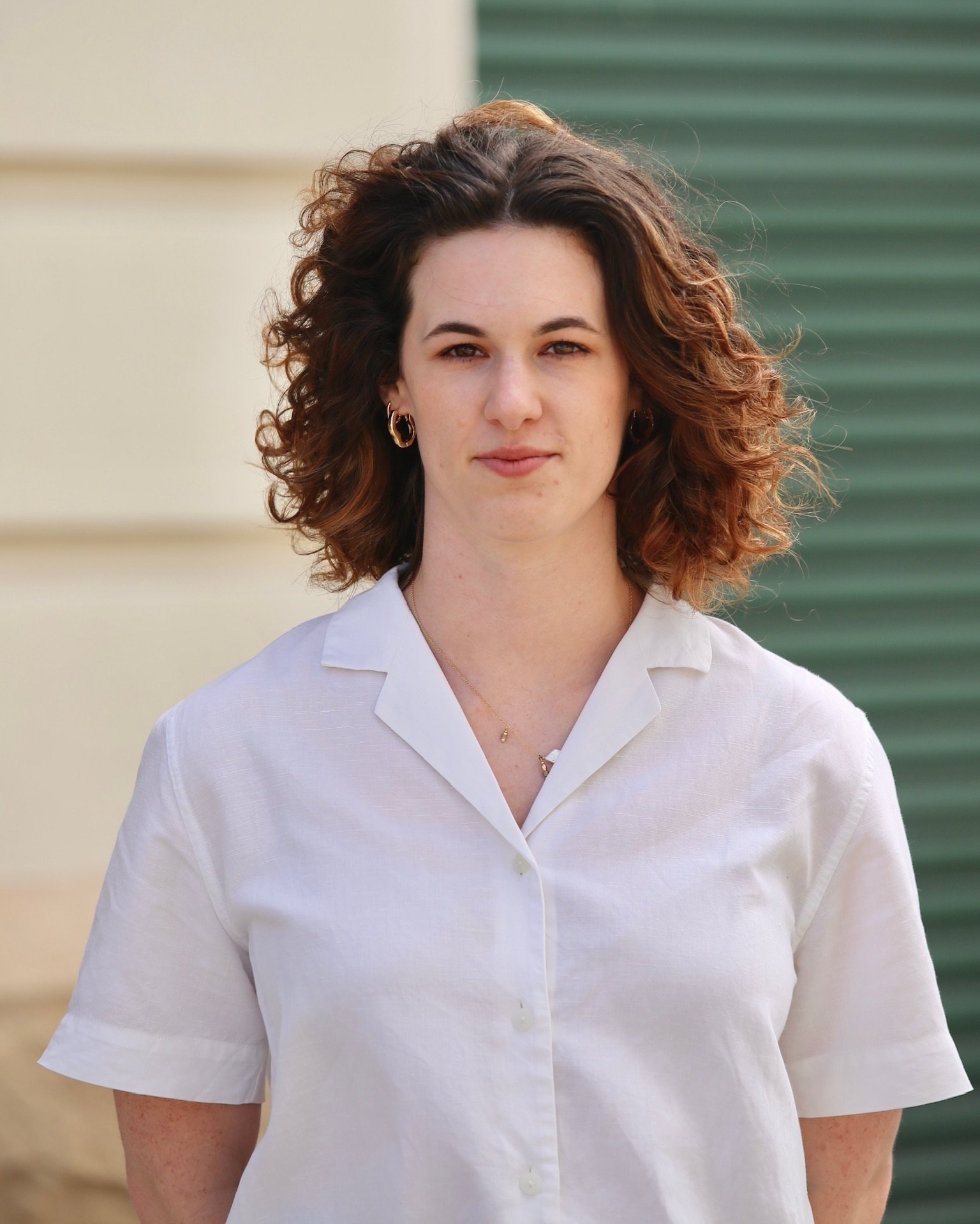 Person with curly hair wearing a white shirt, standing outdoors with a building and green wall in the background.