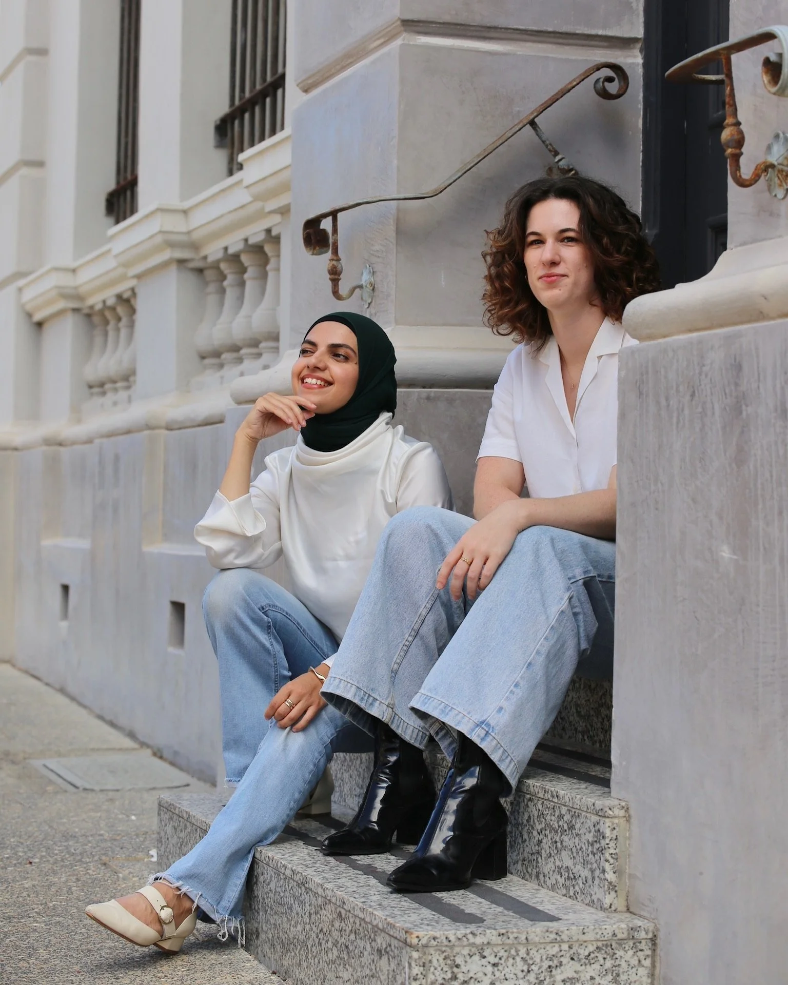 Two women sitting on steps outside a building, one wearing a white blouse and black boots, the other in a black hijab and beige shoes, both smiling.