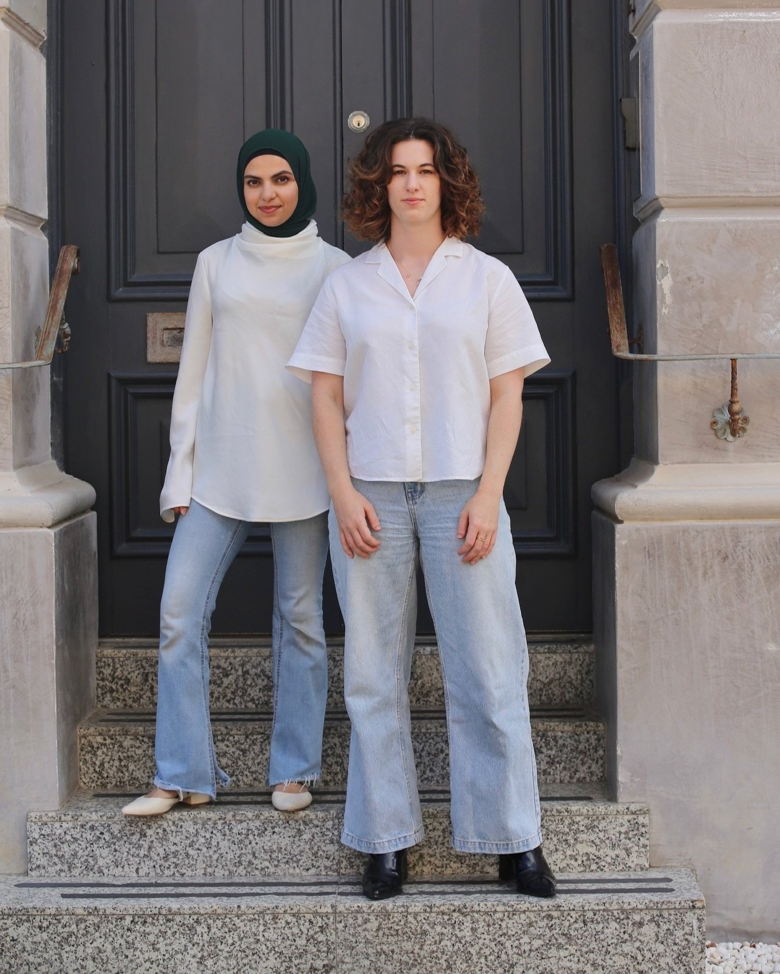 Two women standing on steps in front of a black door. One is wearing a white top, green hijab, and blue jeans. The other is in a white short-sleeved shirt and light blue jeans.