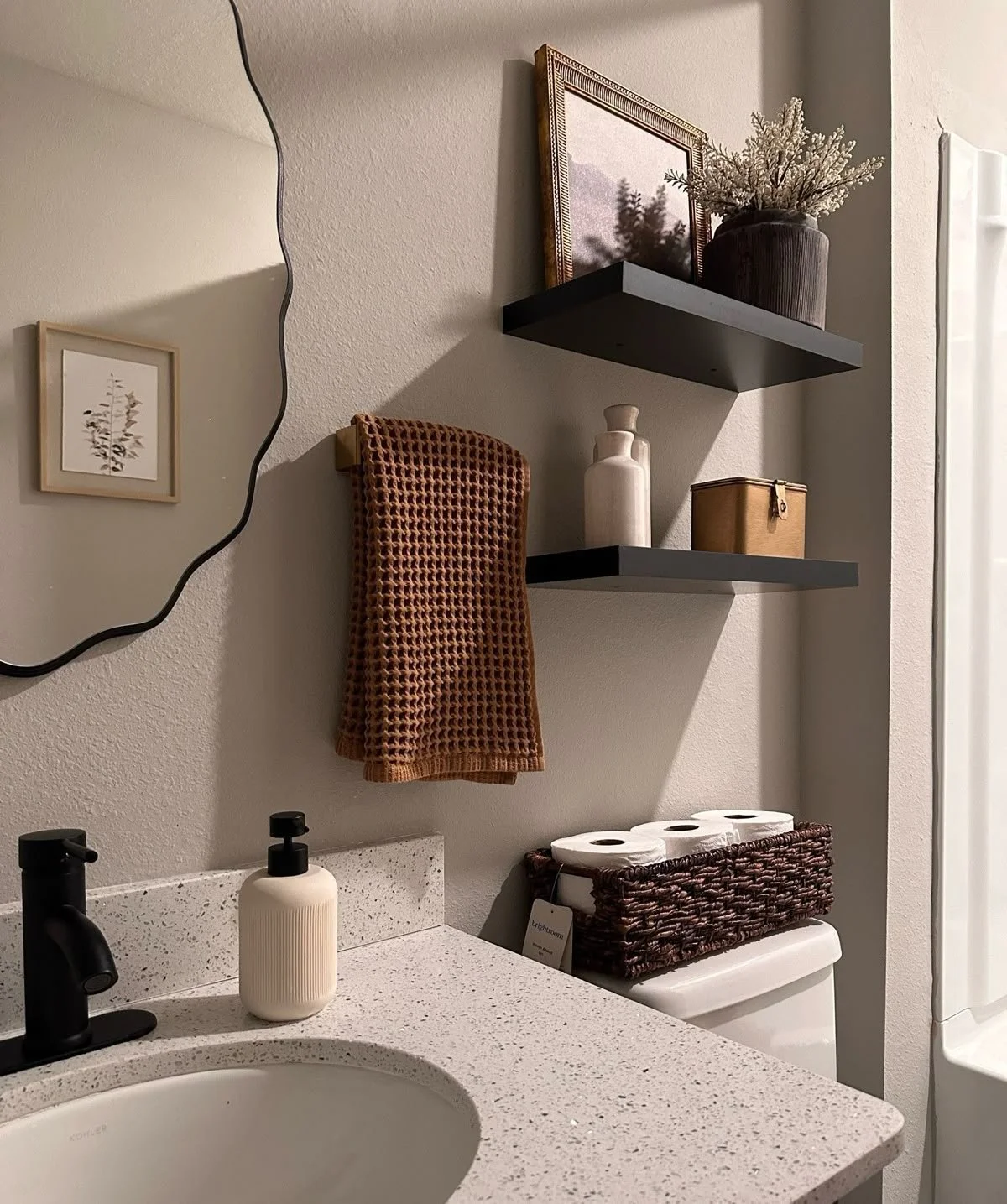 Bathroom scene with a counter featuring a sink, black faucet, and soap dispenser. Above the counter, there are two black shelves with decorative items like a picture frame, vase with dried flowers, and a small container. A brown textured towel hangs on a wall hook. A basket holding toilet paper sits on top of the toilet tank.