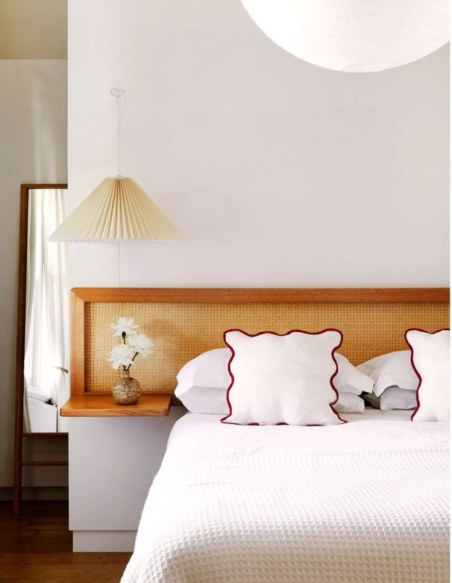 Bedroom with a white bedspread, white pillows with red trim, a wicker headboard, a table lamp with a pleated shade, a vase with white flowers, and a wooden floor.