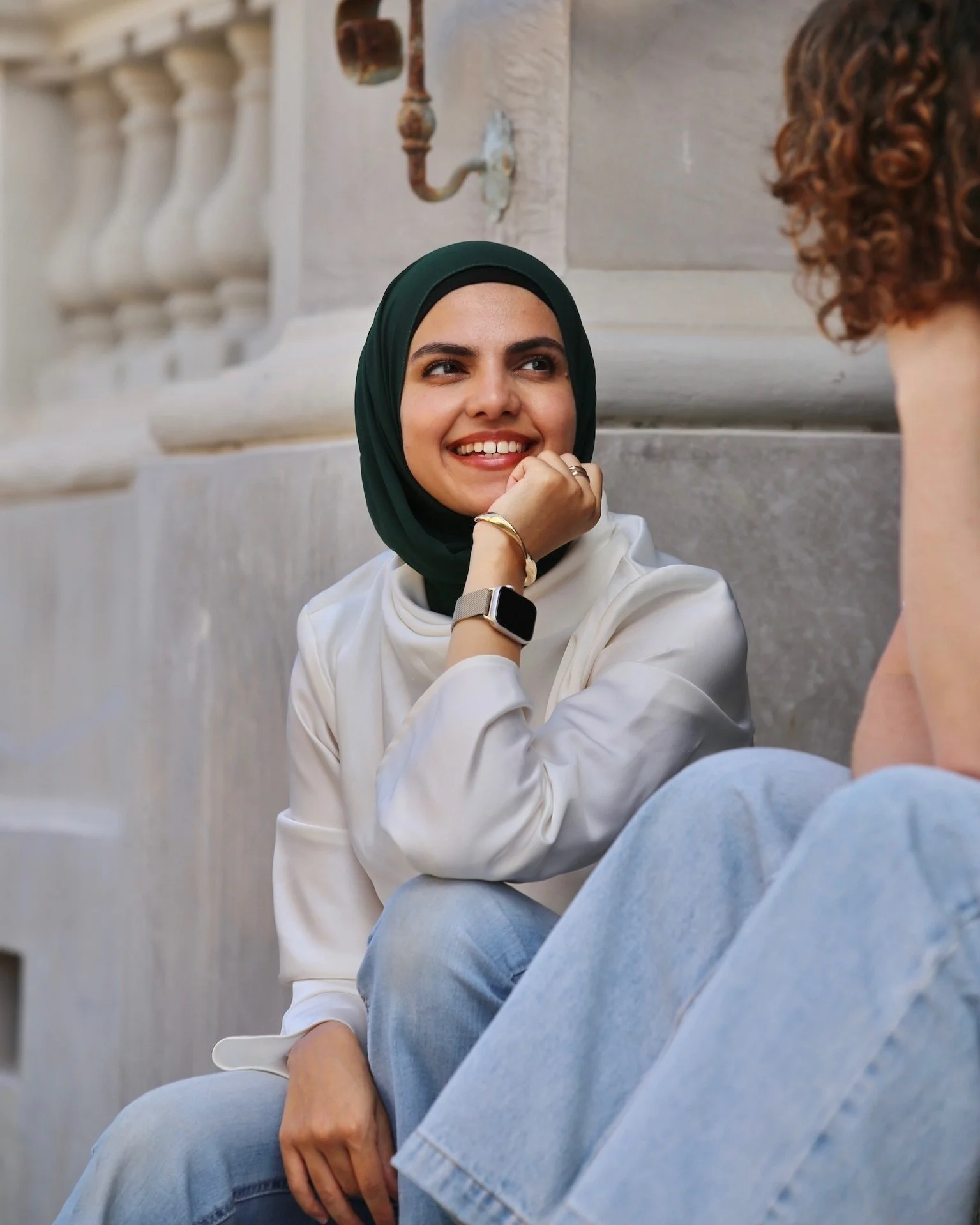 Two people sitting outdoors, one wearing a green hijab and white shirt, smiling, looking at the other person.
